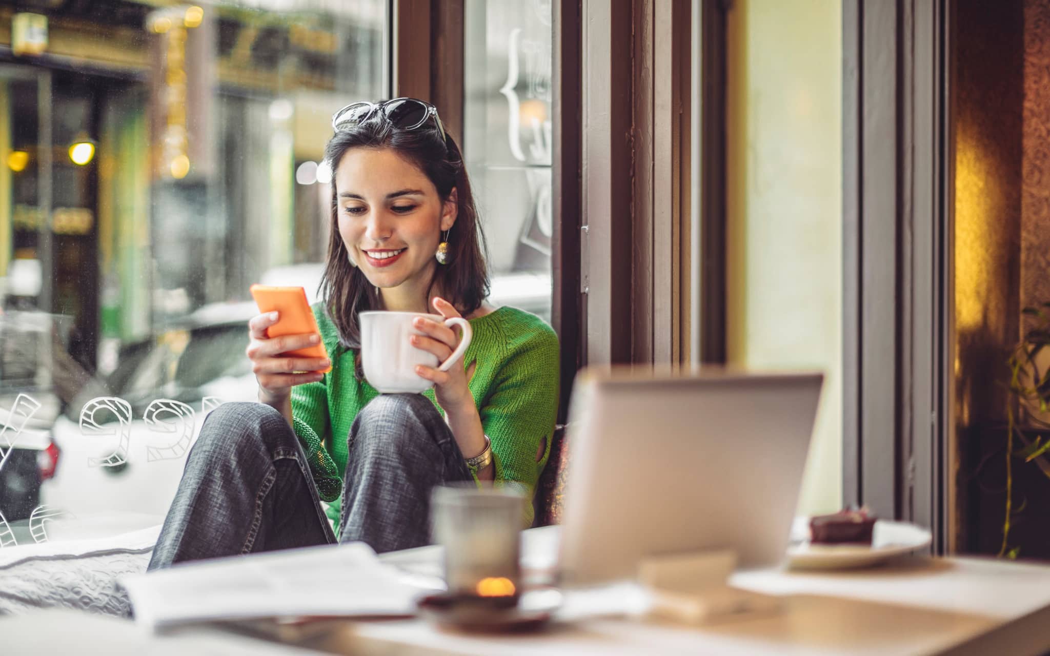 Frau sitzt im Café, guckt lachend in ihr Smartphone und trinkt aus einer Tasse.