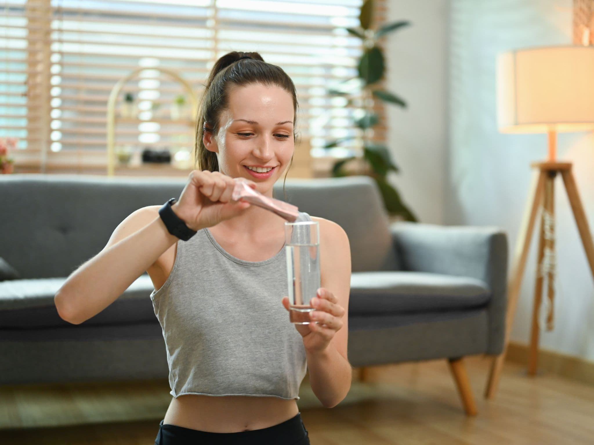 Kollagen: Eine junge Frau sitzt mit einem Glas Wasser vorm Sofa und gibt Kollagenpulver in ihr Getränk.