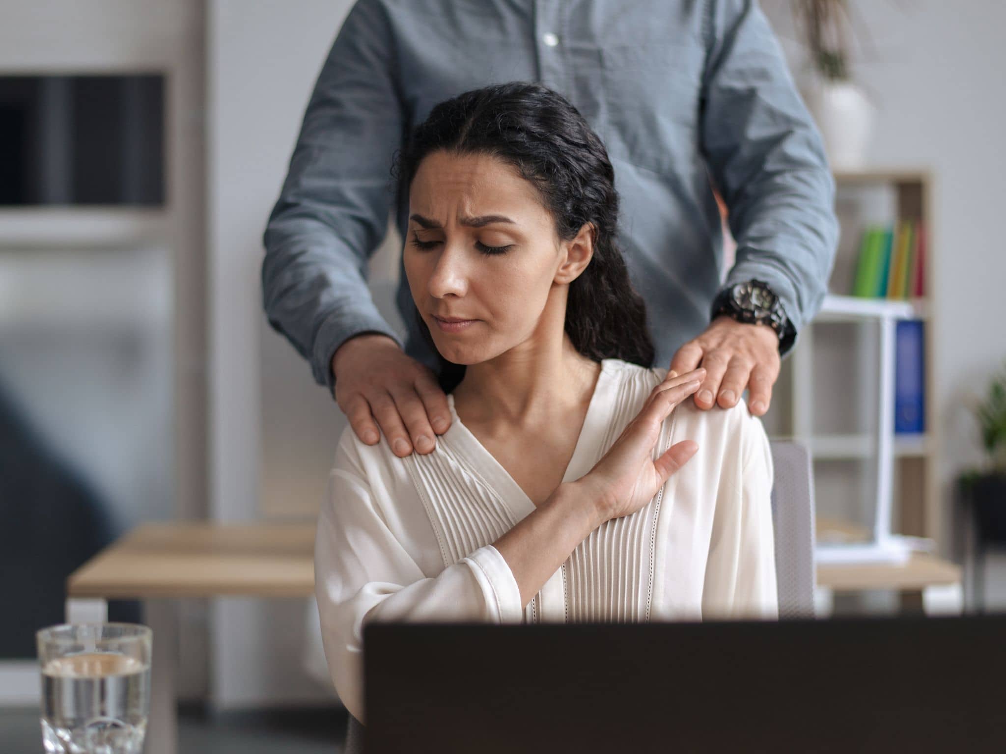 Wann beginnt sexuelle Belästigung: Eine Frau, die am Schreibtisch sitzt, wird von einem Mann an den Schultern berührt.