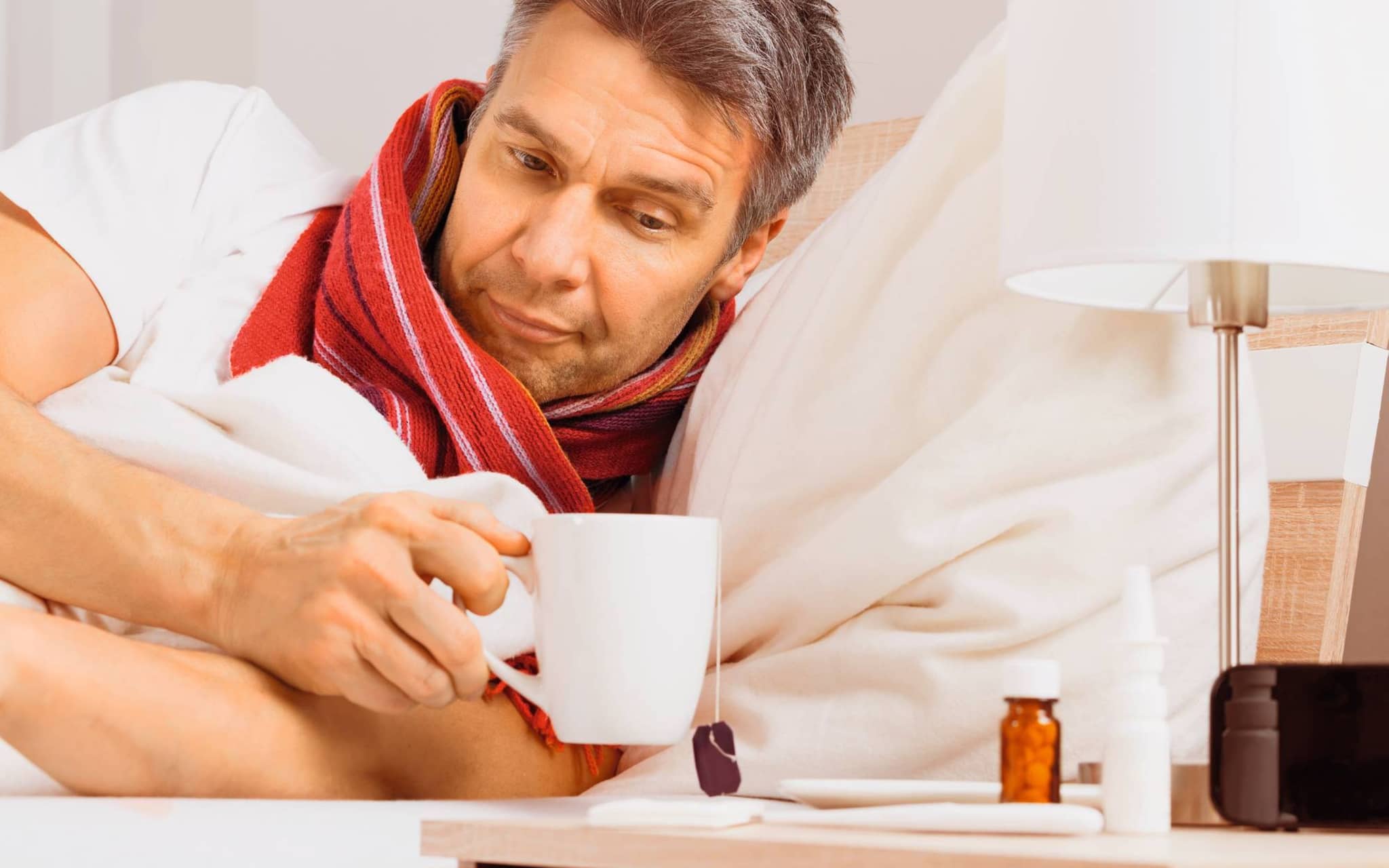 Ein Mann liegt mit Schal im Bett und trinkt Tee - ein bewährtes Hausmittel gegen Halsschmerzen