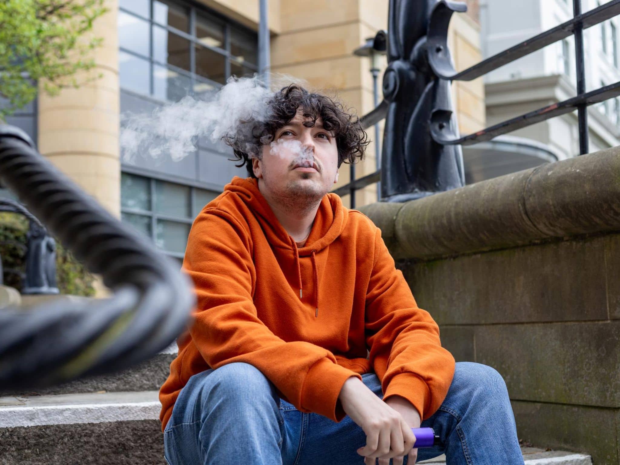Junger Mann sitzt auf einer Treppe und lässt das Nikotin seiner E-Zigarette auf sich wirken.