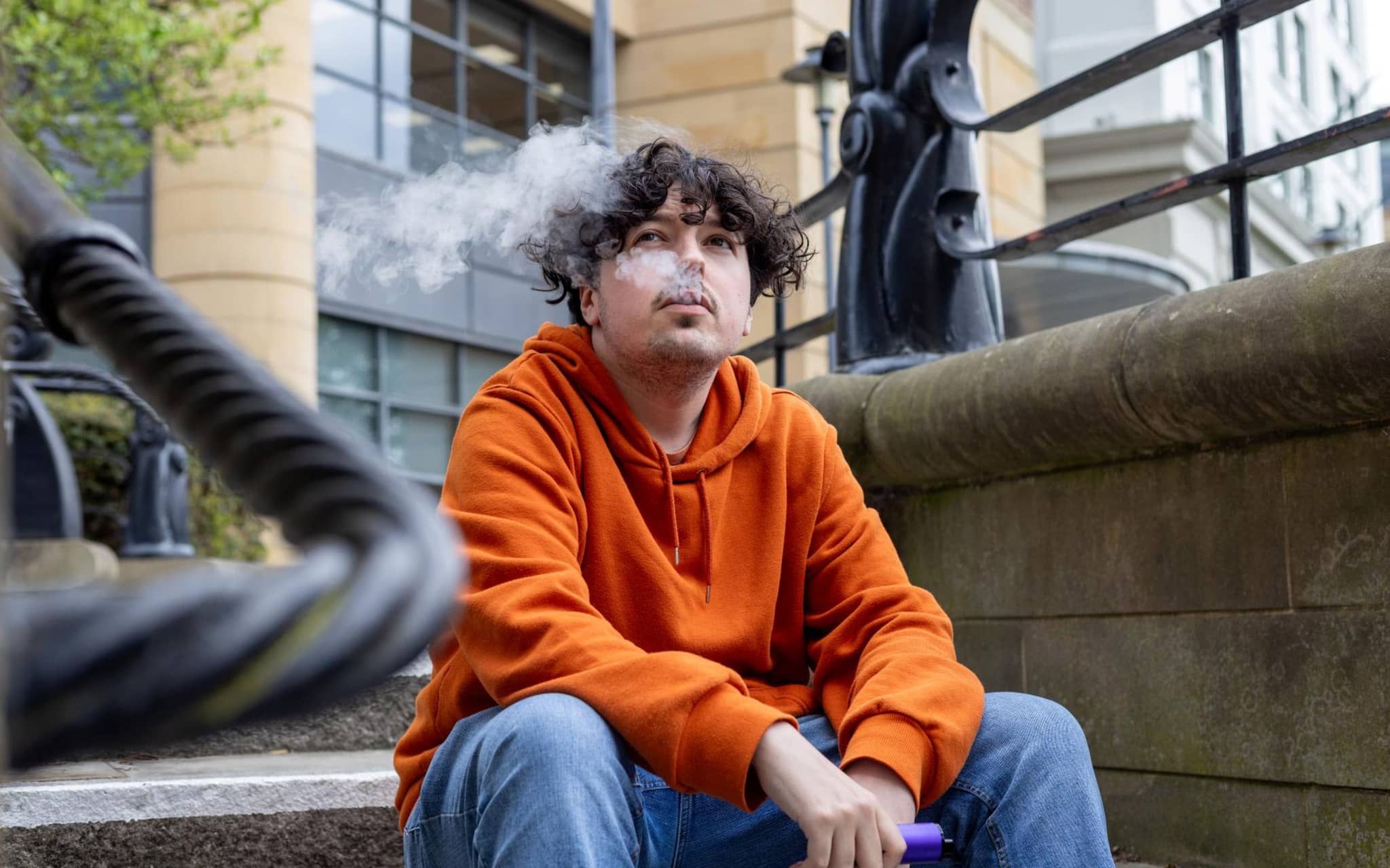 Junger Mann sitzt auf einer Treppe und lässt das Nikotin seiner E-Zigarette auf sich wirken.