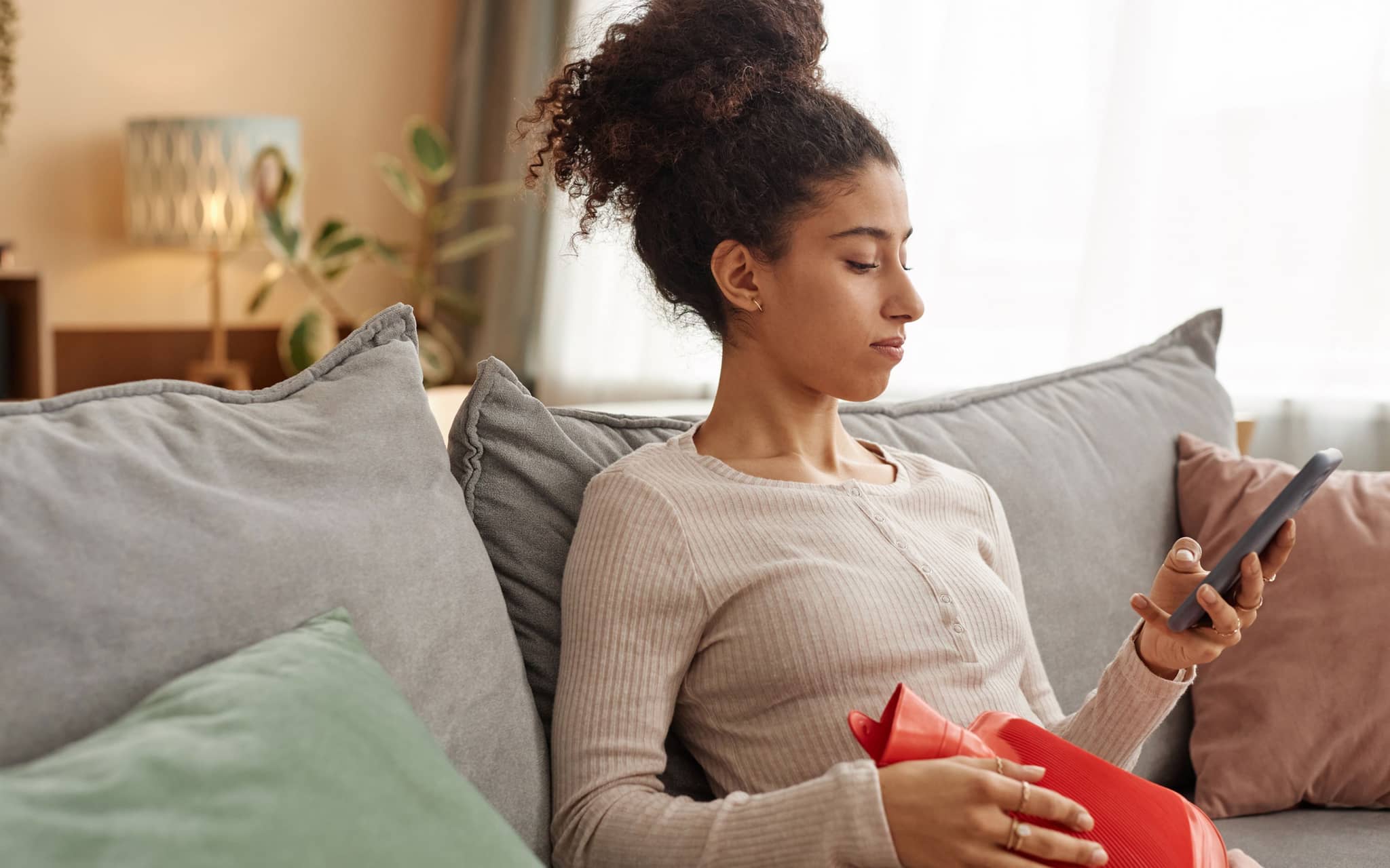 Eine junge Frau sitzt mit Wärmflasche auf einem Sofa und guckt auf ihr Smartphone