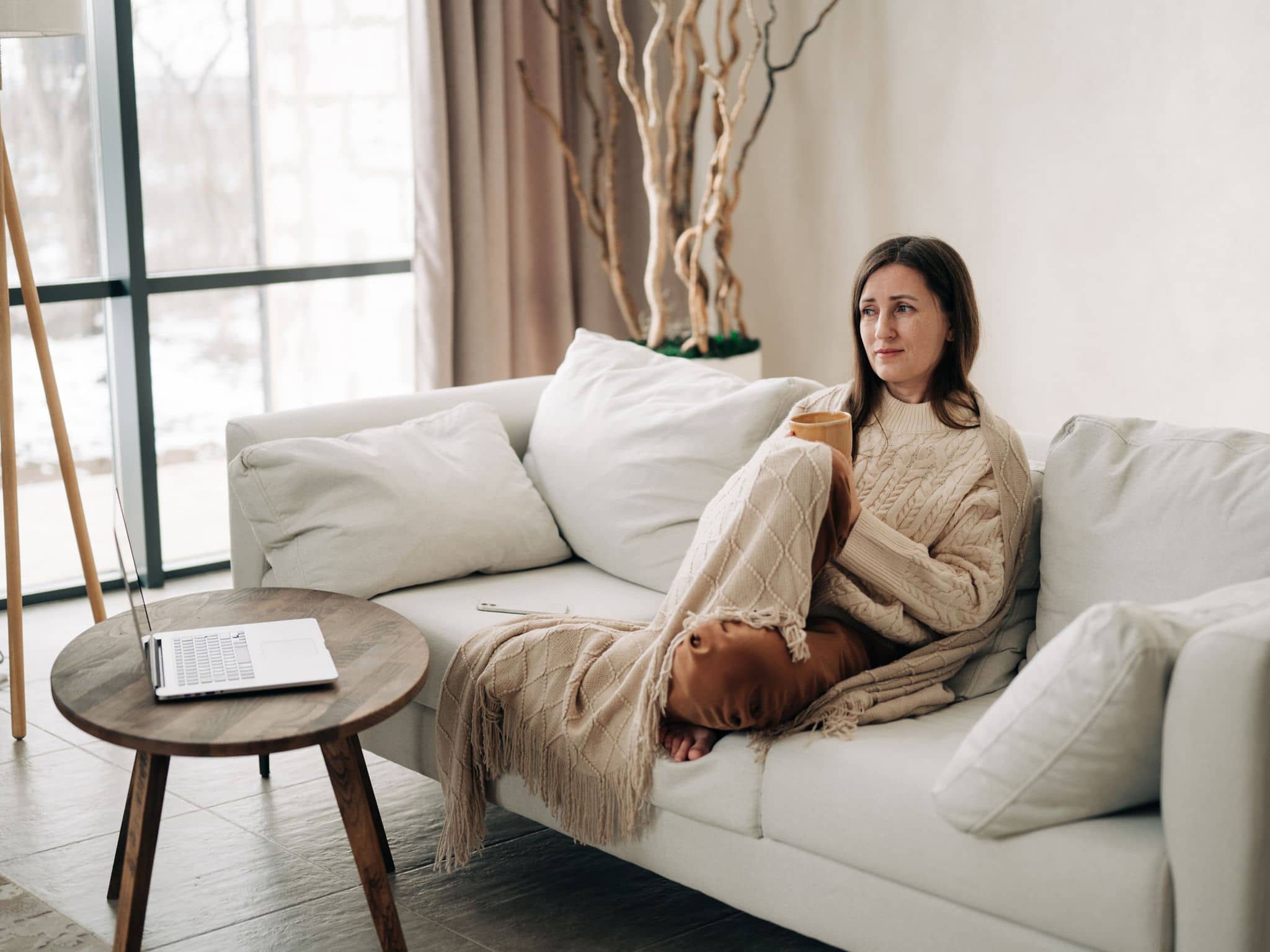Eine Frau sitzt in eine Decke gekuschelt und einem Sofa und denkt nach