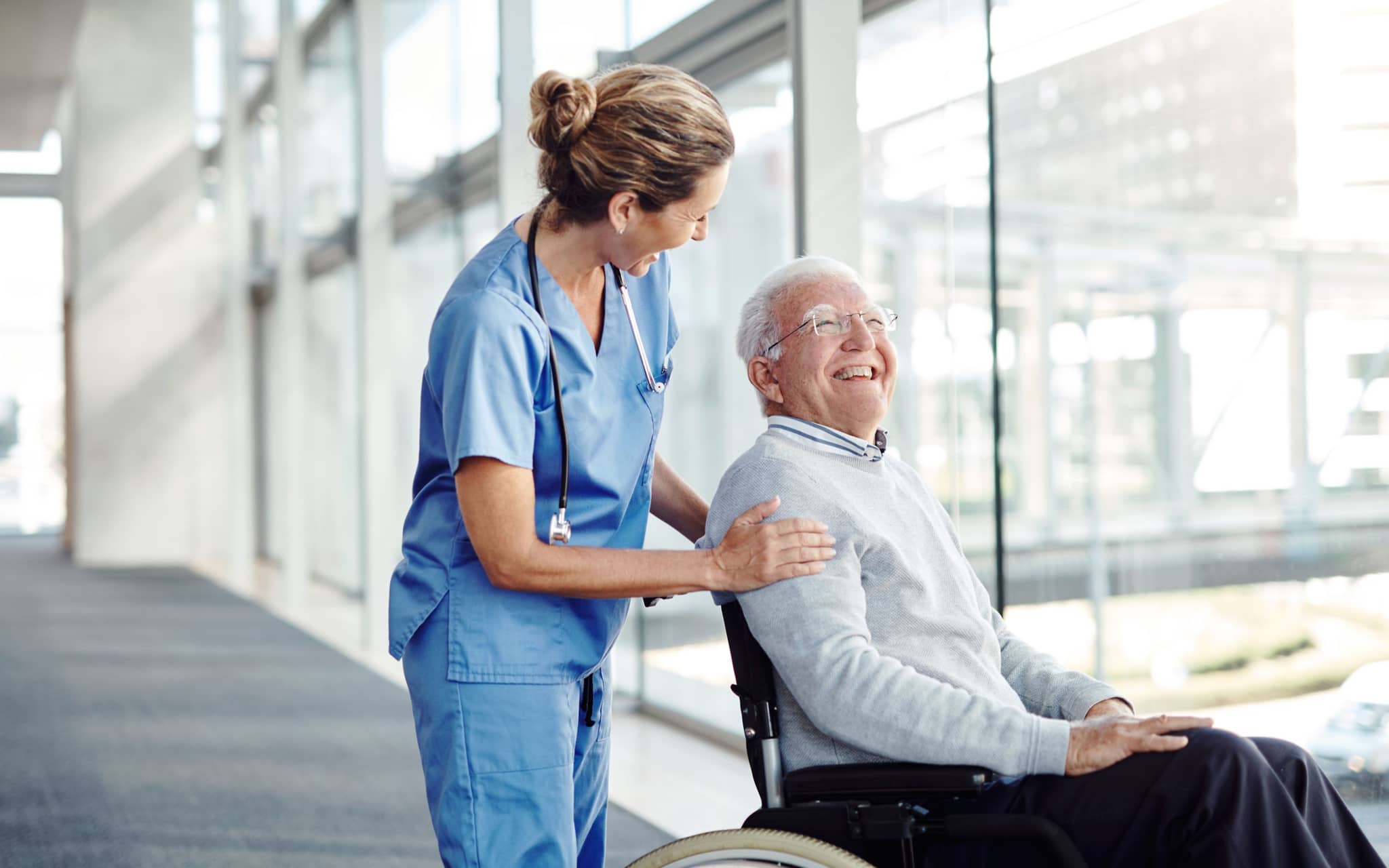 Ein älterer Mann im Rollstuhl wird von einer Pflegerin über einen Flur geschoben