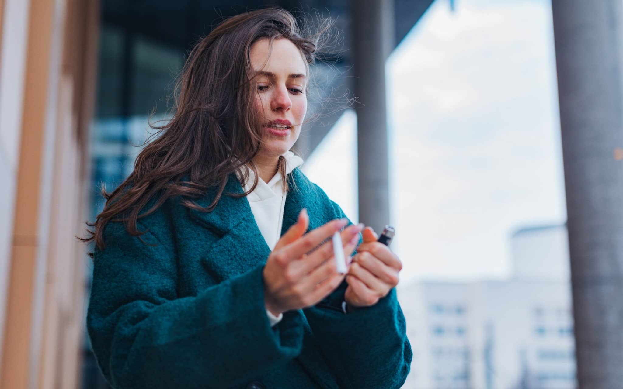 Eine junge Frau steht draußen mit einer Zigarette und einem Feuerzeug in der Hand und guckt auf ihre Hände.