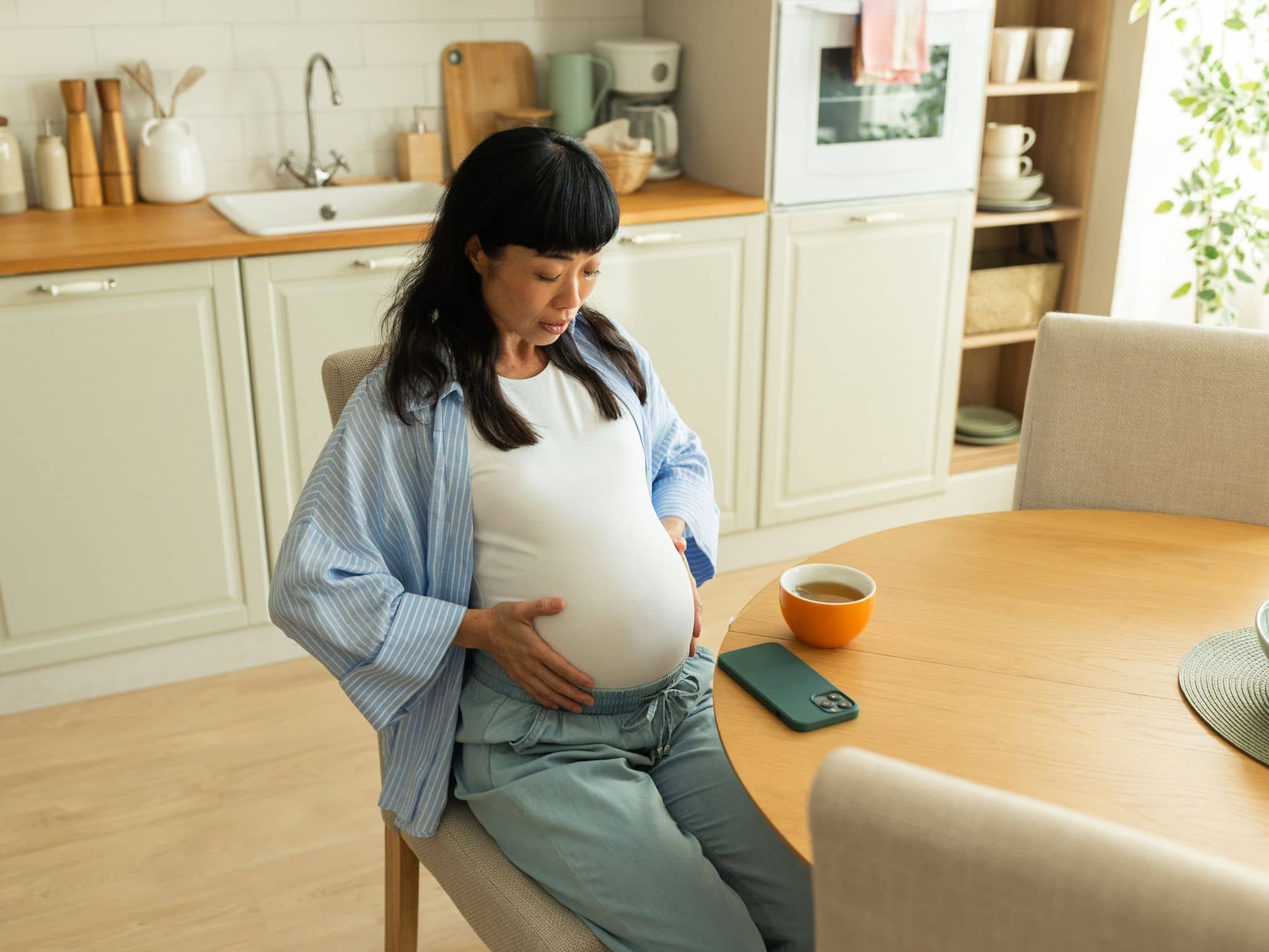 Eine schwangere Frau sitzt auf einem Küchenstuhl und hält sich ihren Bauch.