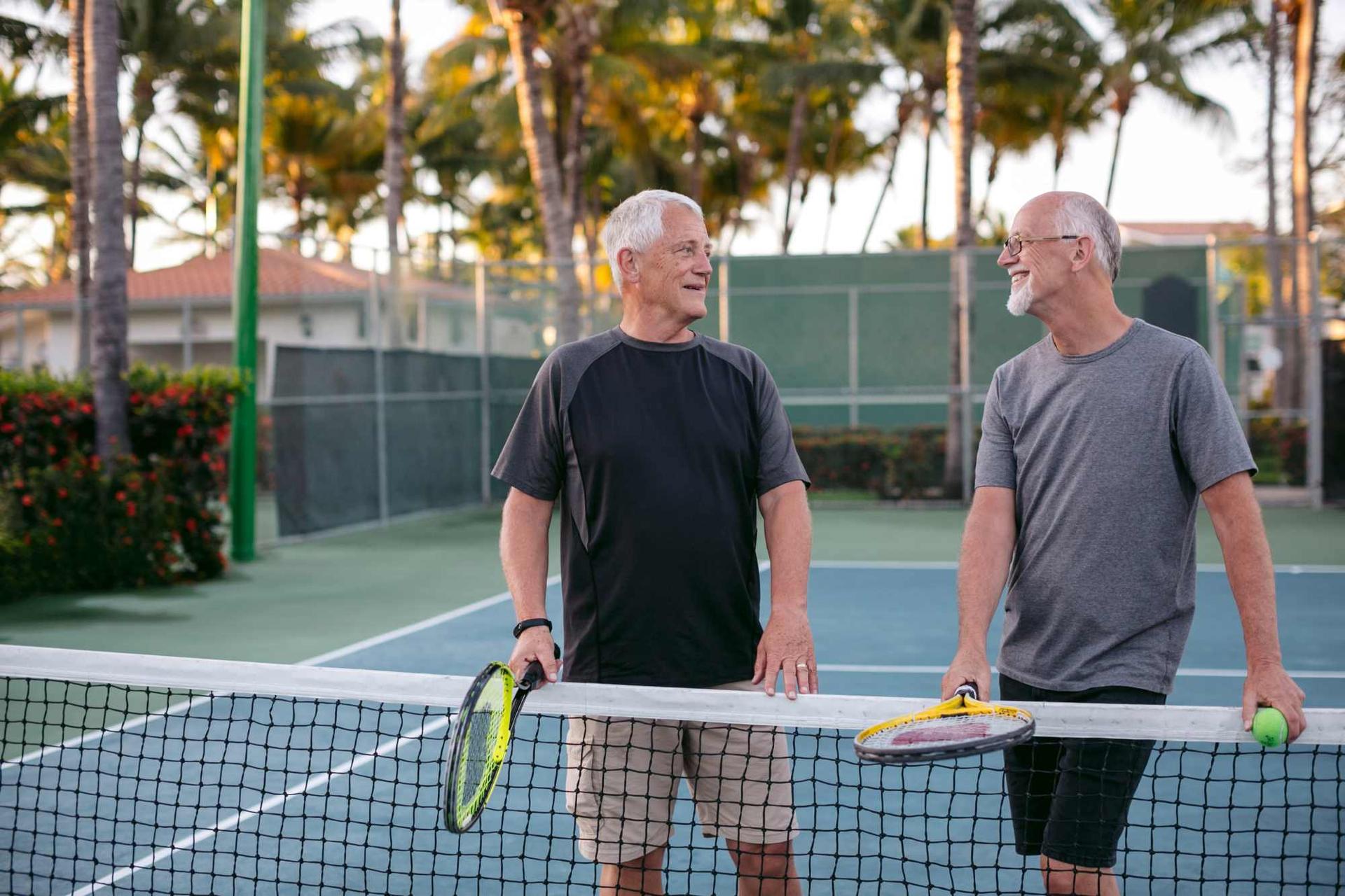 Darmspiegelung: Zwei ältere Männer stehen an einem Tennisnetz, halten Tennisschläger in der Hand und sehen sich an.