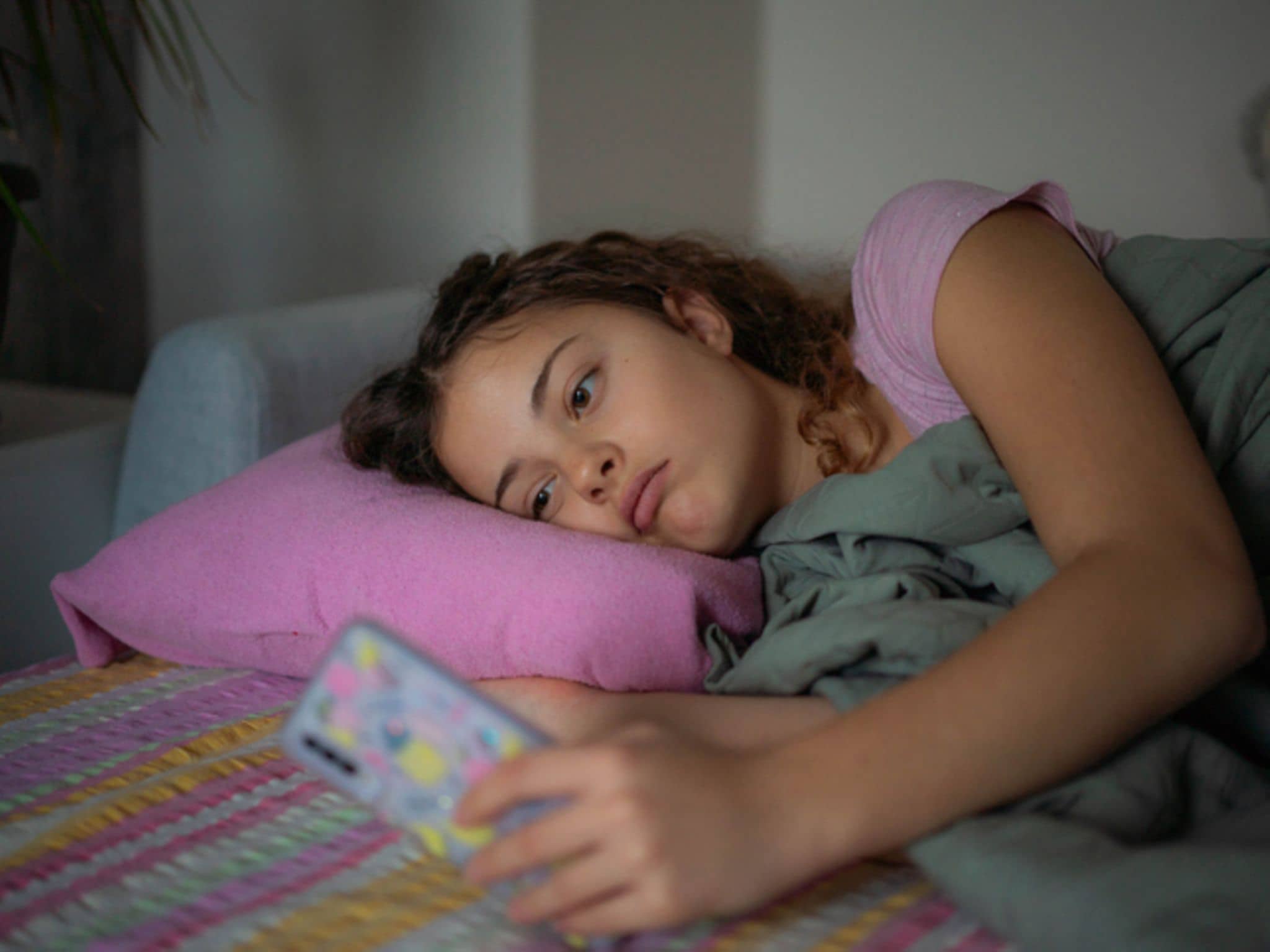 Jugendliche liegt apathisch im Bett und starrt aufs Smartphone.