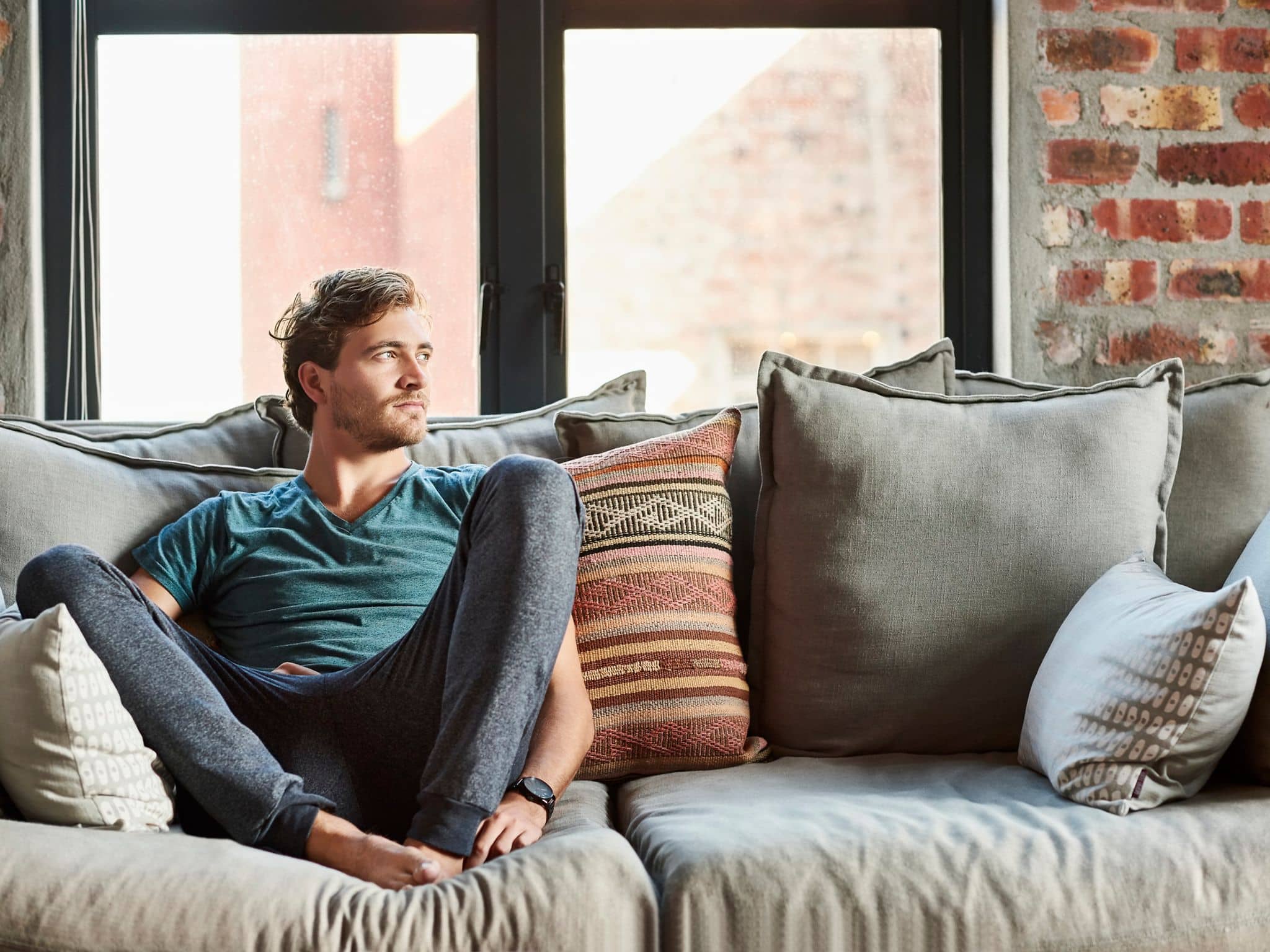Ein junger Mann sitzt mit hochgezogenen Beinen auf einem Sofa und sieht nachdenklich aus.