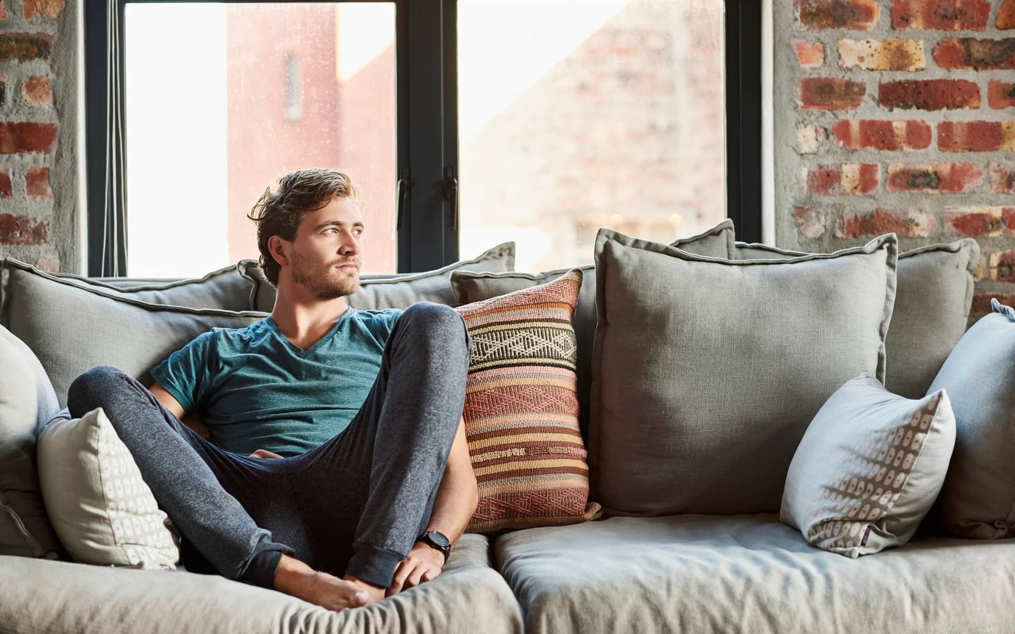 Ein junger Mann sitzt mit hochgezogenen Beinen auf einem Sofa und sieht nachdenklich aus.