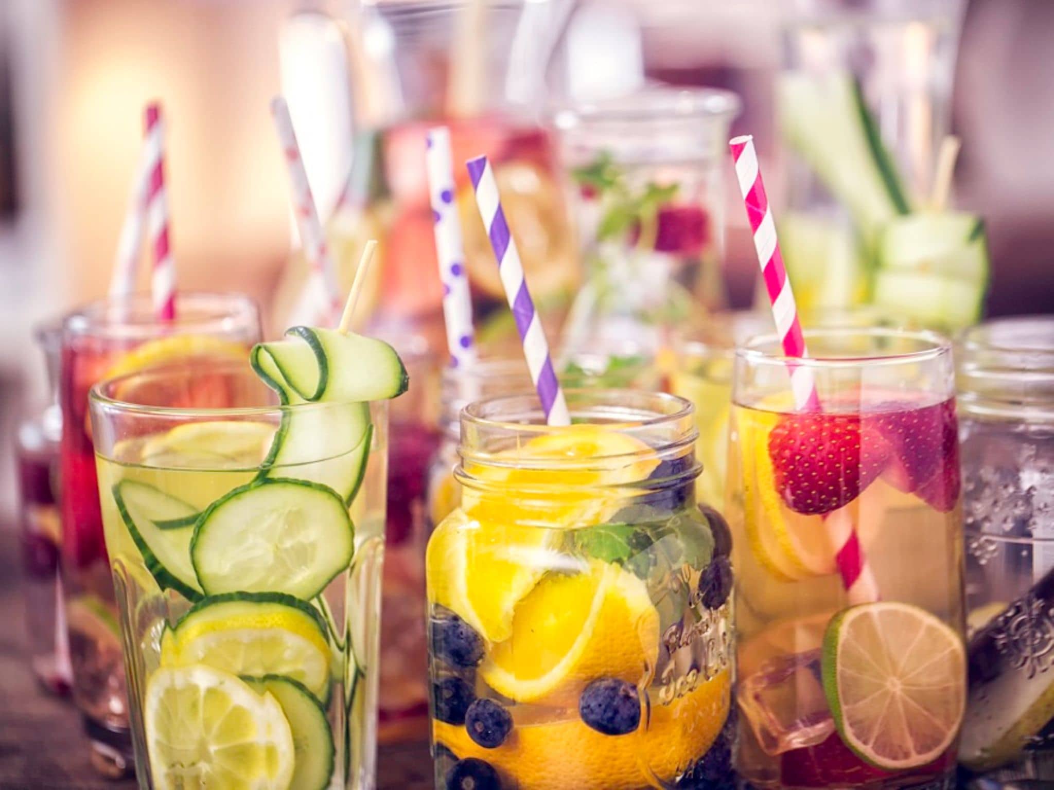 Gläser mit Wasser und frischen Früchten wie Zitrone, Gurke, Beeren und Limette, mit bunten Trinkhalmen.