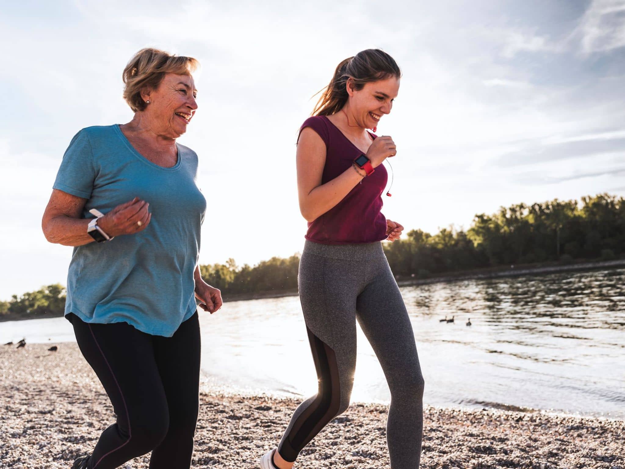 Eine ältere Frau und eine jüngere Frau laufen gemeinsam sportlich an einem Ufer entlang.