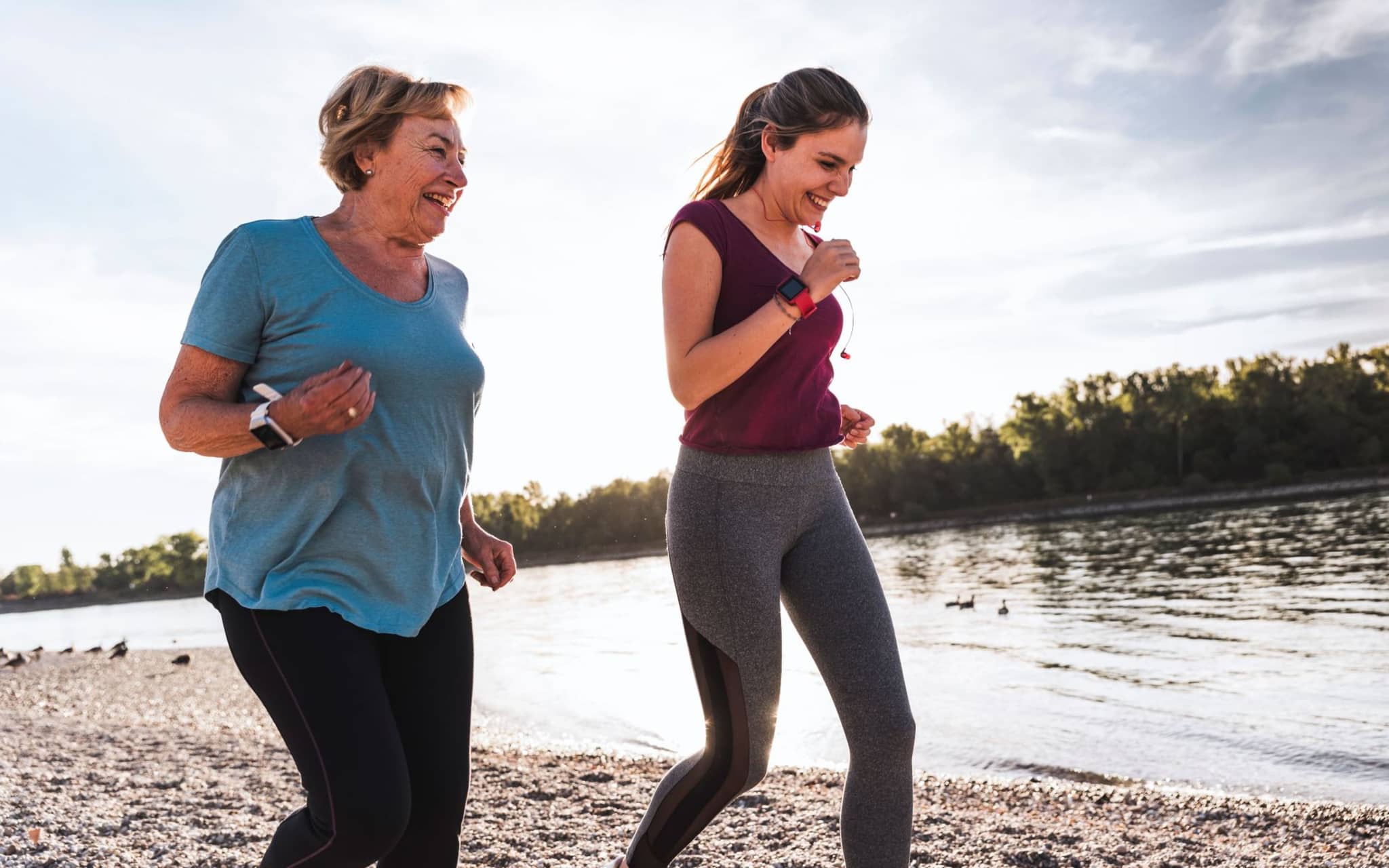 Eine ältere Frau und eine jüngere Frau laufen gemeinsam sportlich an einem Ufer entlang.