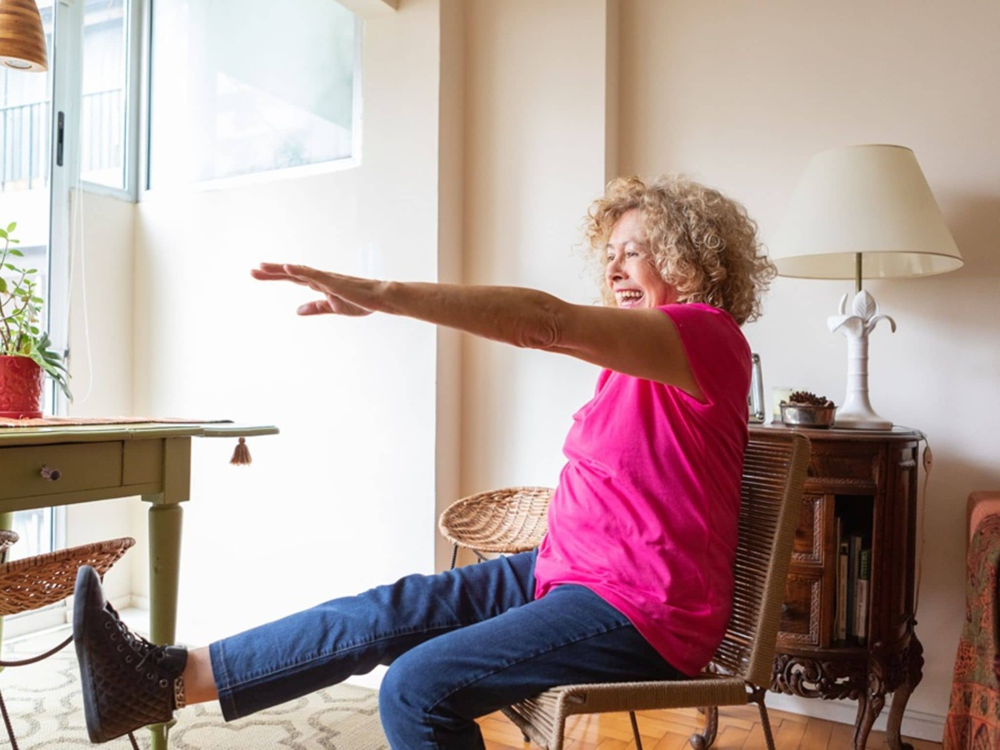 Ältere Frau macht eine Gleichgewichtsübung im Sitzen und hebt ein Bein in ihrem Wohnzimmer.