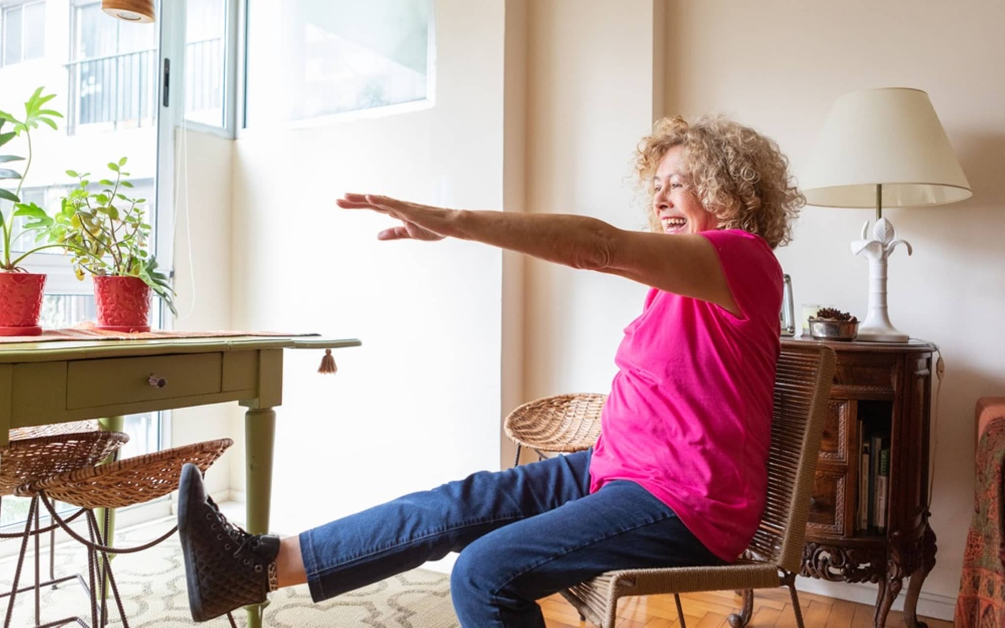 Ältere Frau macht eine Gleichgewichtsübung im Sitzen und hebt ein Bein in ihrem Wohnzimmer.