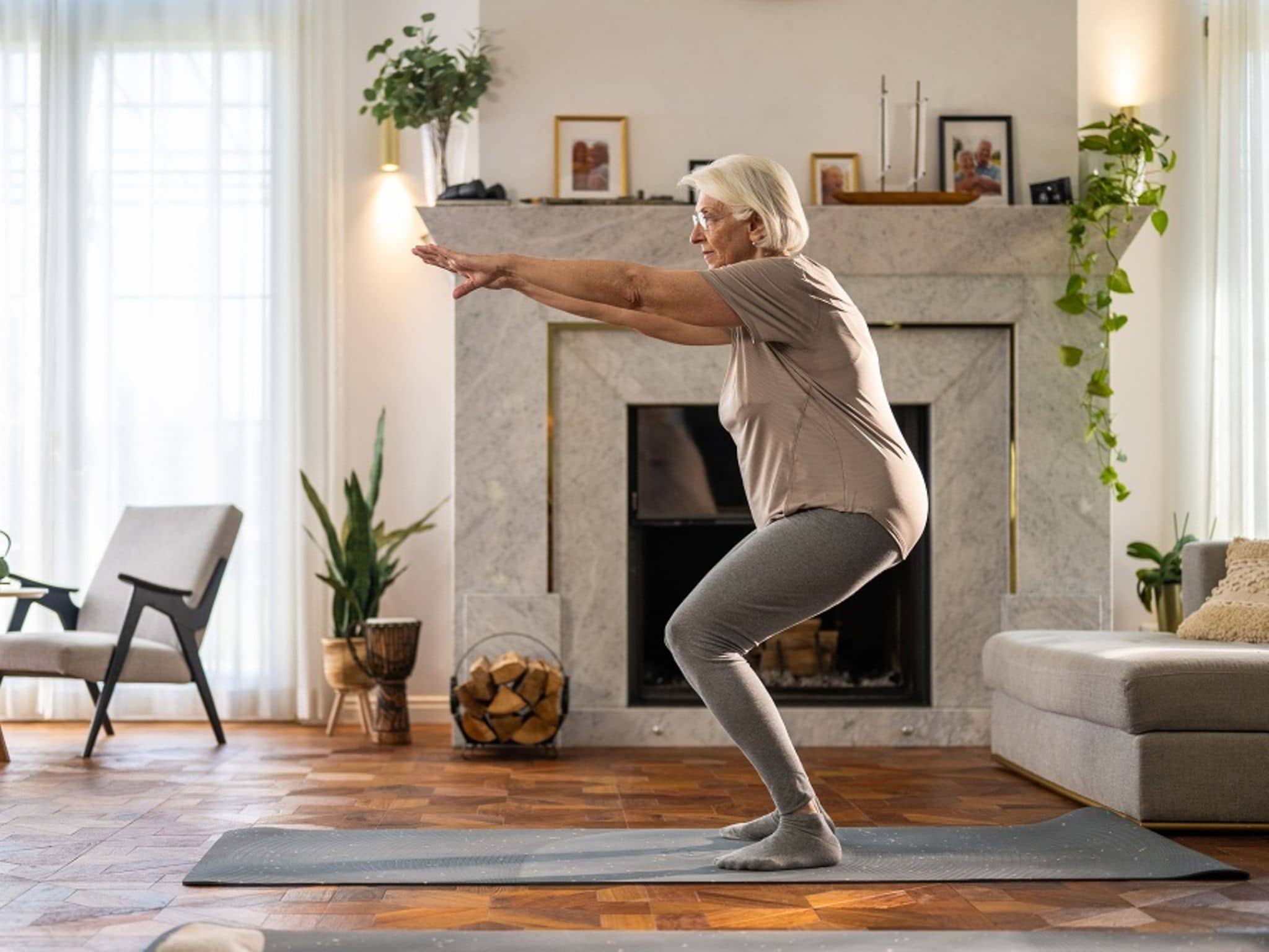 Ältere Frau macht eine Kniebeuge auf einer Yogamatte im Wohnzimmer.