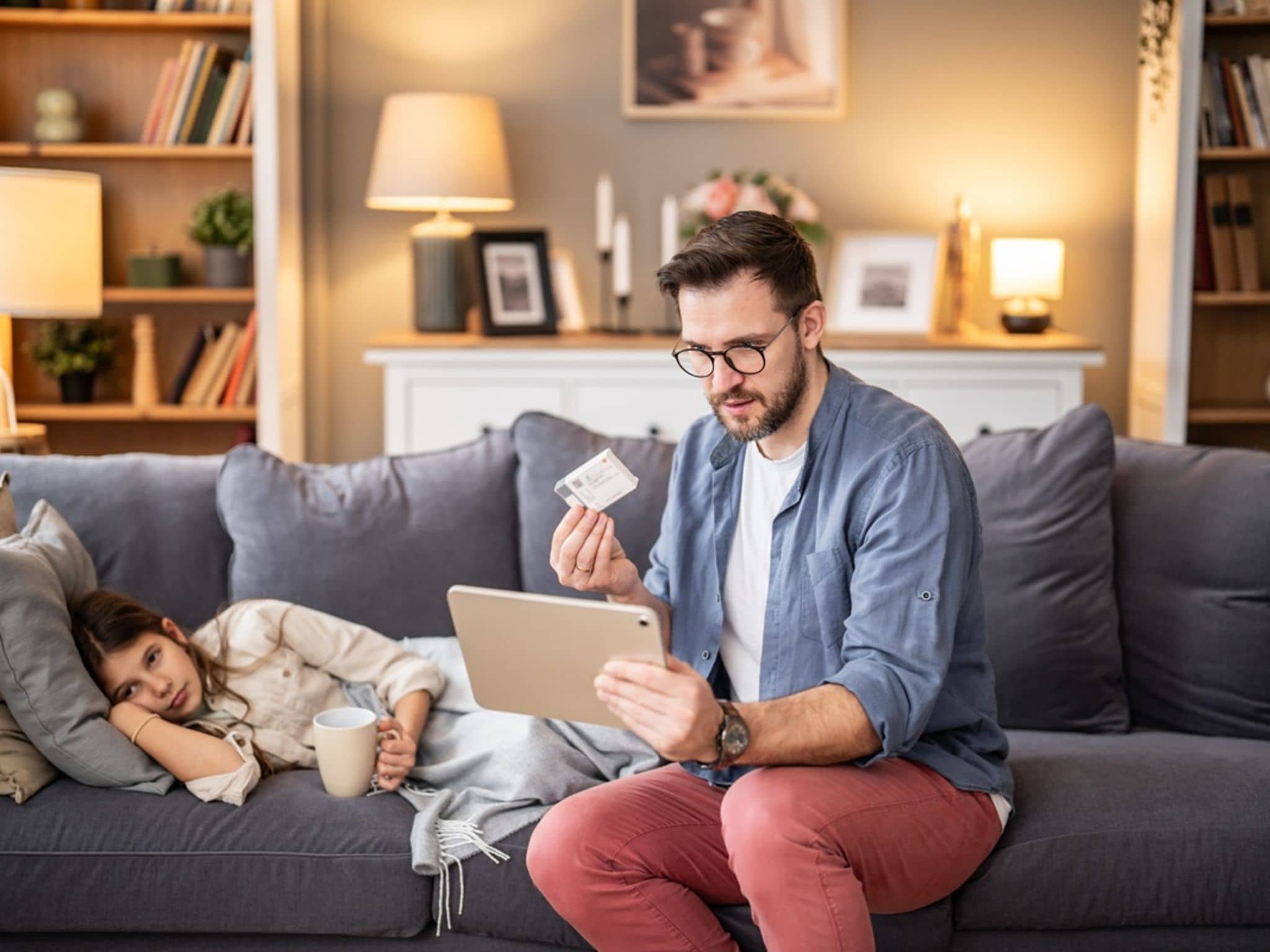 Mann sitzt mit Tablet und Karte auf dem Sofa, neben ihm liegt ein krank wirkendes Kind mit Decke und Tasse.