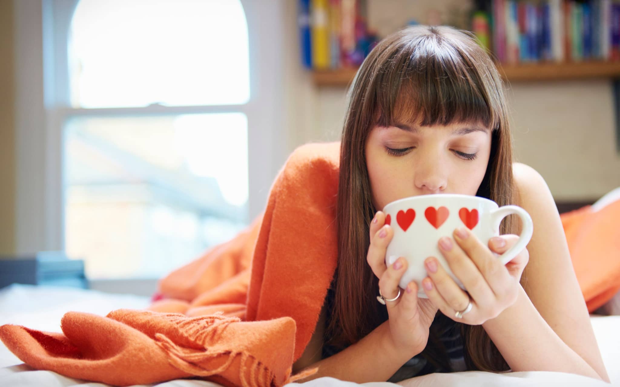 Eine junge Frau liegt mit dem Bauch auf dem Bett und genießt einen Becher Tee.