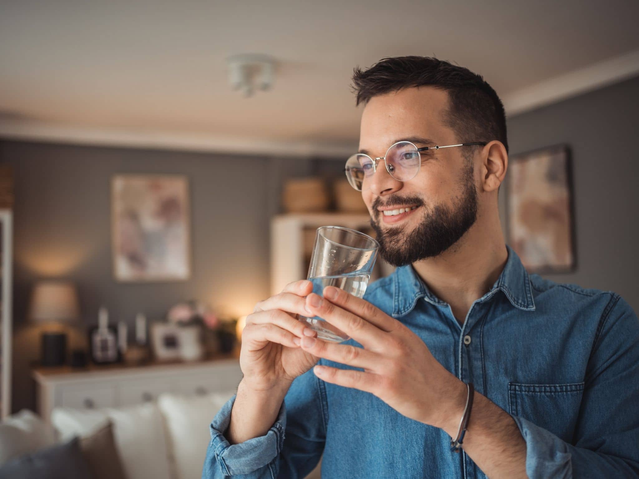 Lächelnder Mann mit Brille hält ein Glas Wasser in den Händen und steht in einem gemütlichen Wohnzimmer.