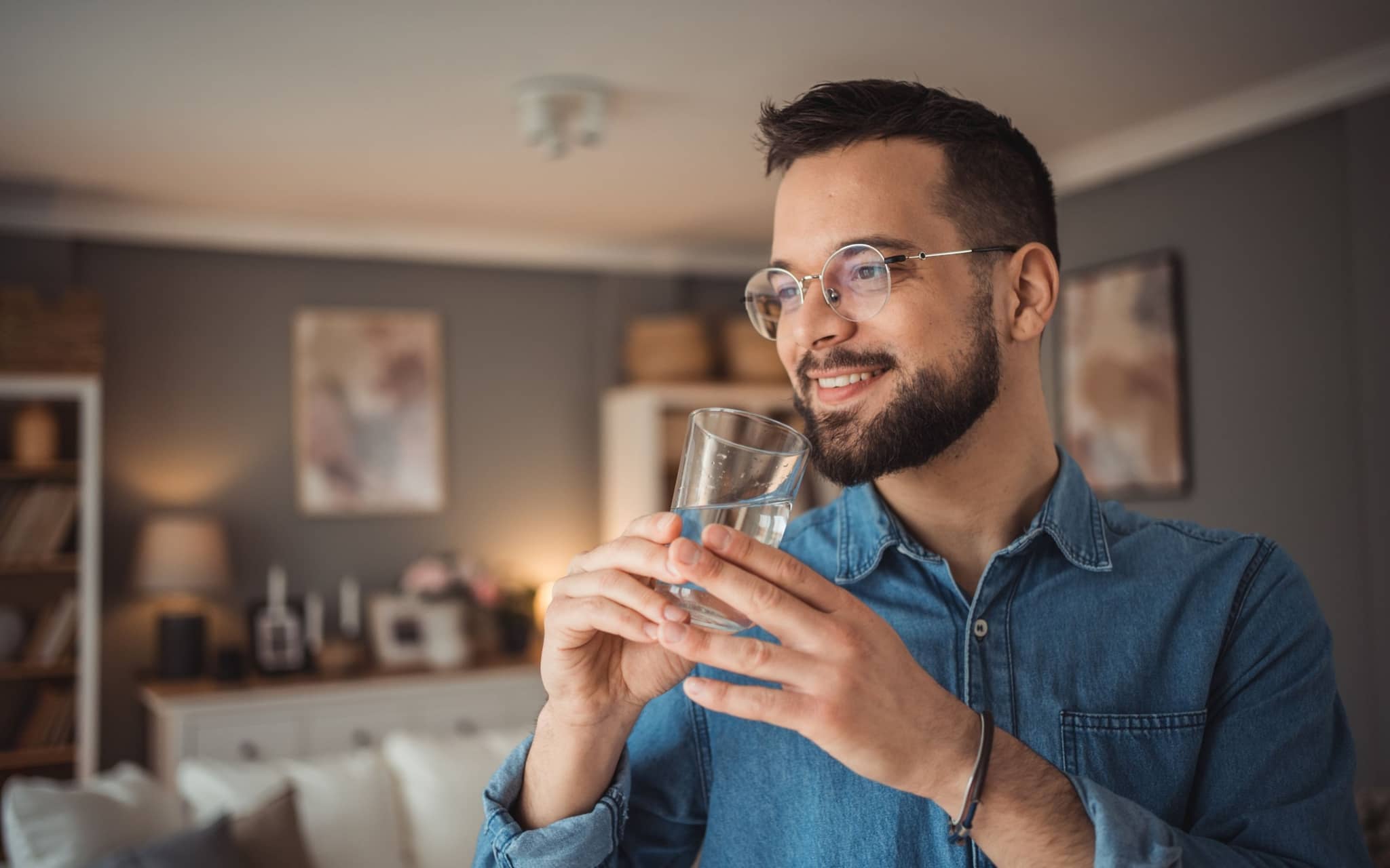 Lächelnder Mann mit Brille hält ein Glas Wasser in den Händen und steht in einem gemütlichen Wohnzimmer.