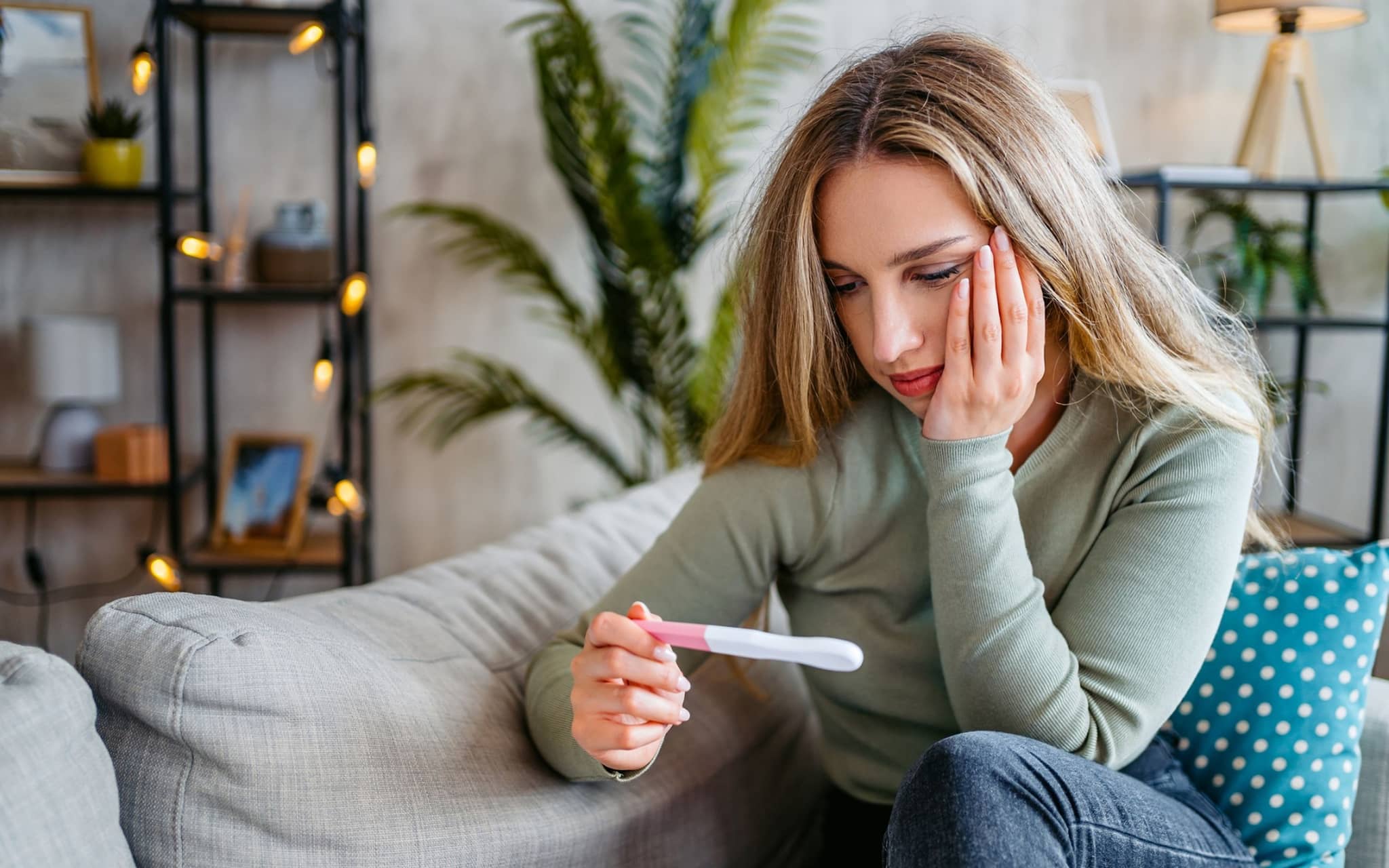 Junge Frau sitzt auf dem Sofa, hält einen Schwangerschaftstest in der Hand und schaut nachdenklich darauf.
