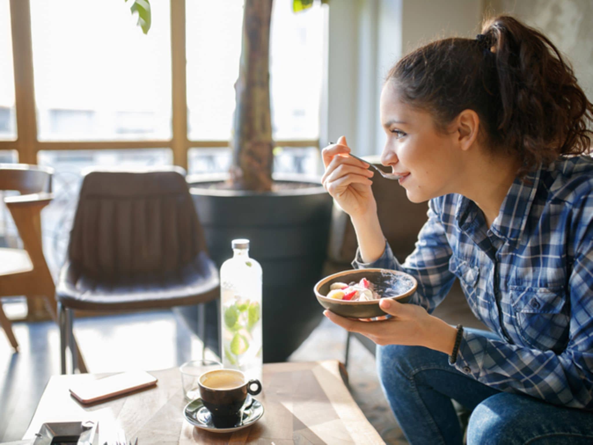 Milchersatzprodukte: Eine junge Frau sitzt an einem Tisch und isst Müsli mit Früchten aus einer Schale