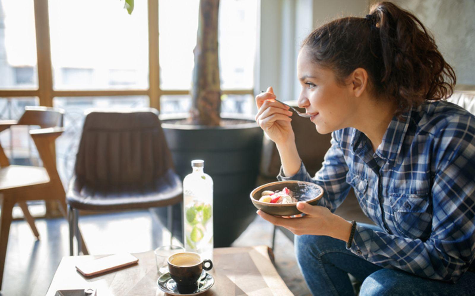 Milchersatzprodukte: Eine junge Frau sitzt an einem Tisch und isst Müsli mit Früchten aus einer Schale
