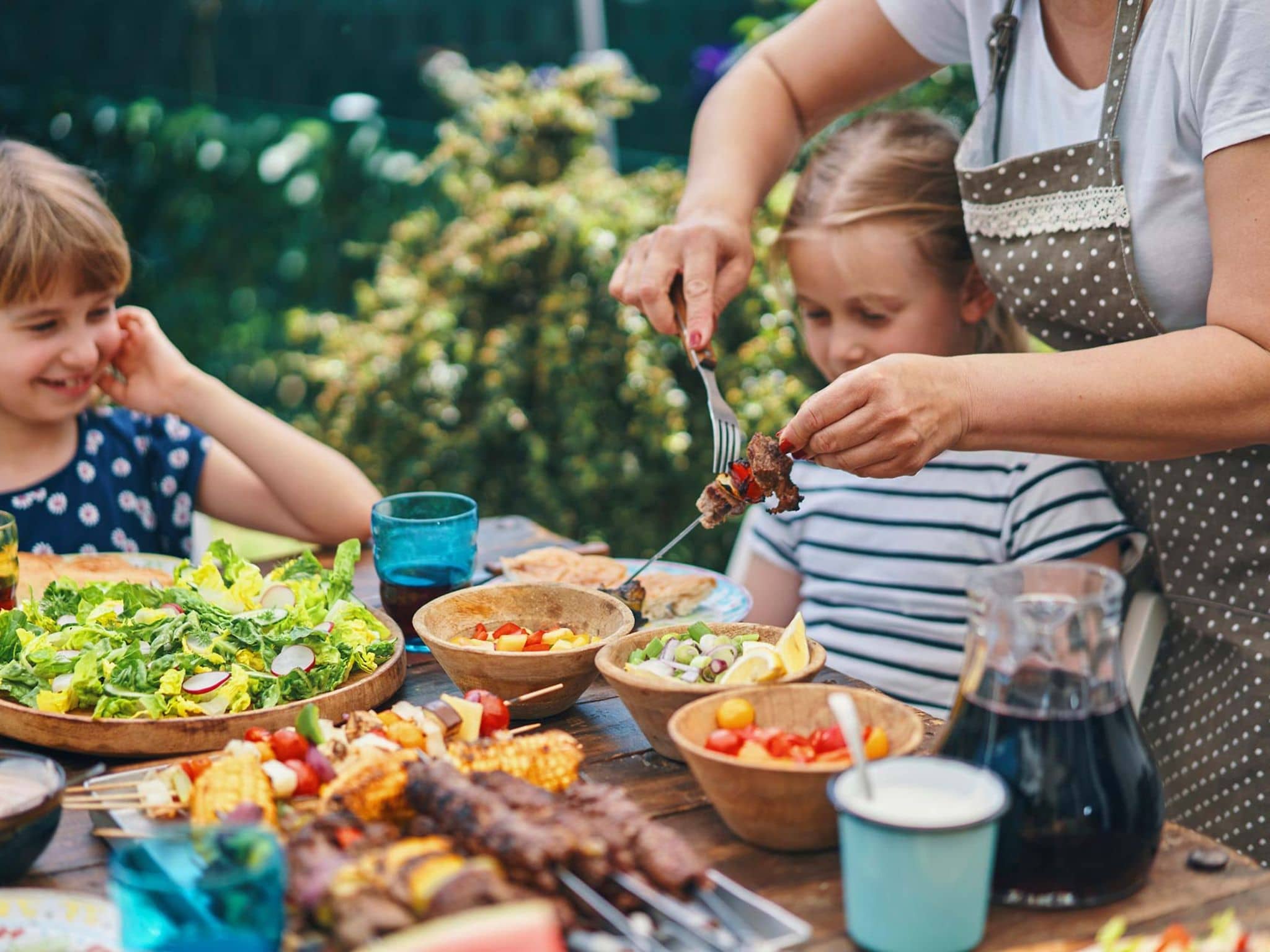 Gesund grillen: Zwei Kinder sitzen draußen an einem gedeckten Tisch und bekommen von der Mutter Grillfleisch auf den Teller.