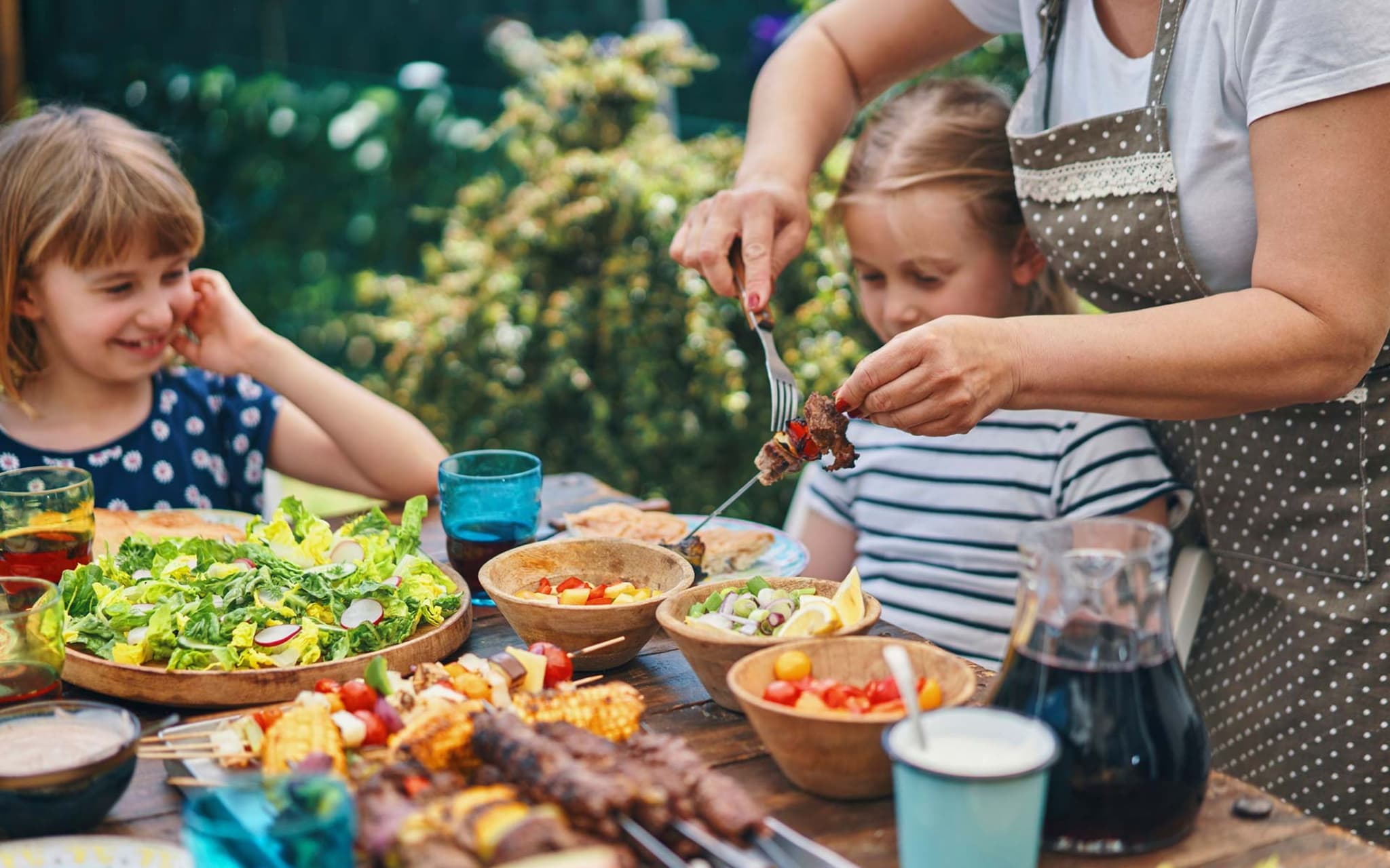 Gesund grillen: Zwei Kinder sitzen draußen an einem gedeckten Tisch und bekommen von der Mutter Grillfleisch auf den Teller.