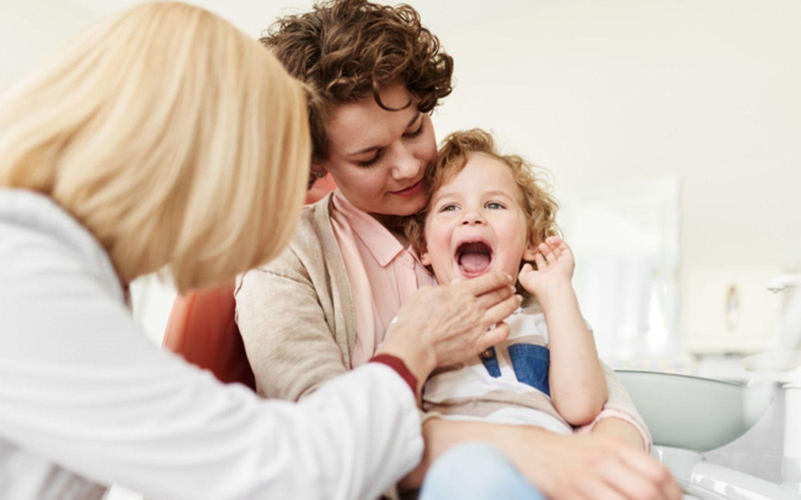 Symbolbild für Angst vorm Zahnarzt: Eine Mutter hat ihr Kind auf den Schoß und die Zahnärztin schaut in den Mund des Kindes