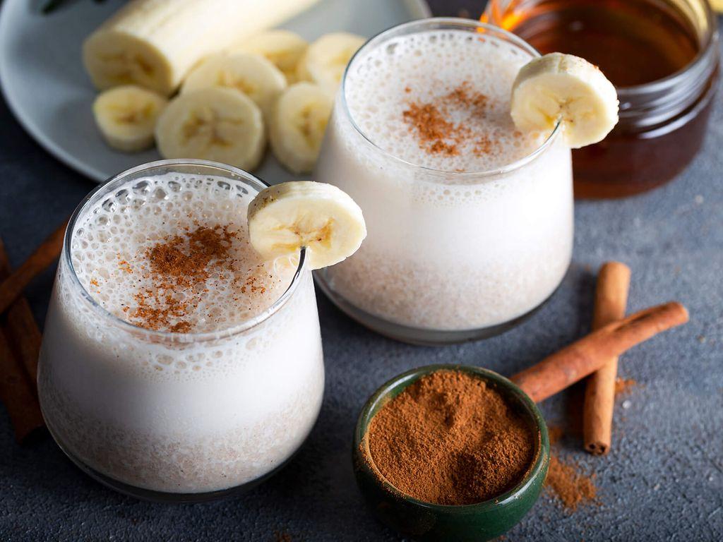 Rezept Bananenmilch: Bild von zwei Gläsern mit Bananenmilch, daneben Bananen, Honig und Zimt.