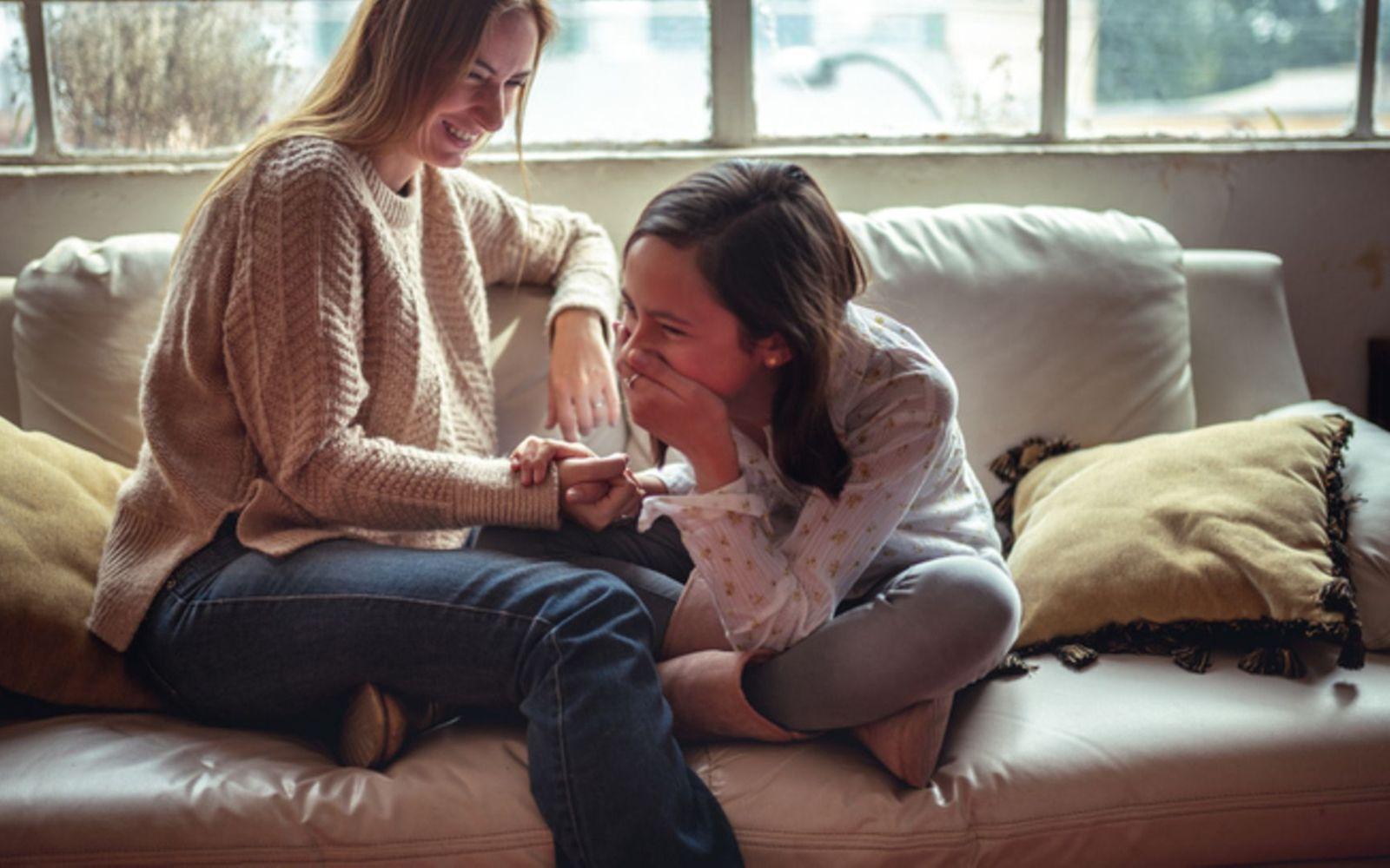 Mutter und Tochter sitzen vertraut auf der Couch und die Tochter lacht mit der Hand vor dem Mund