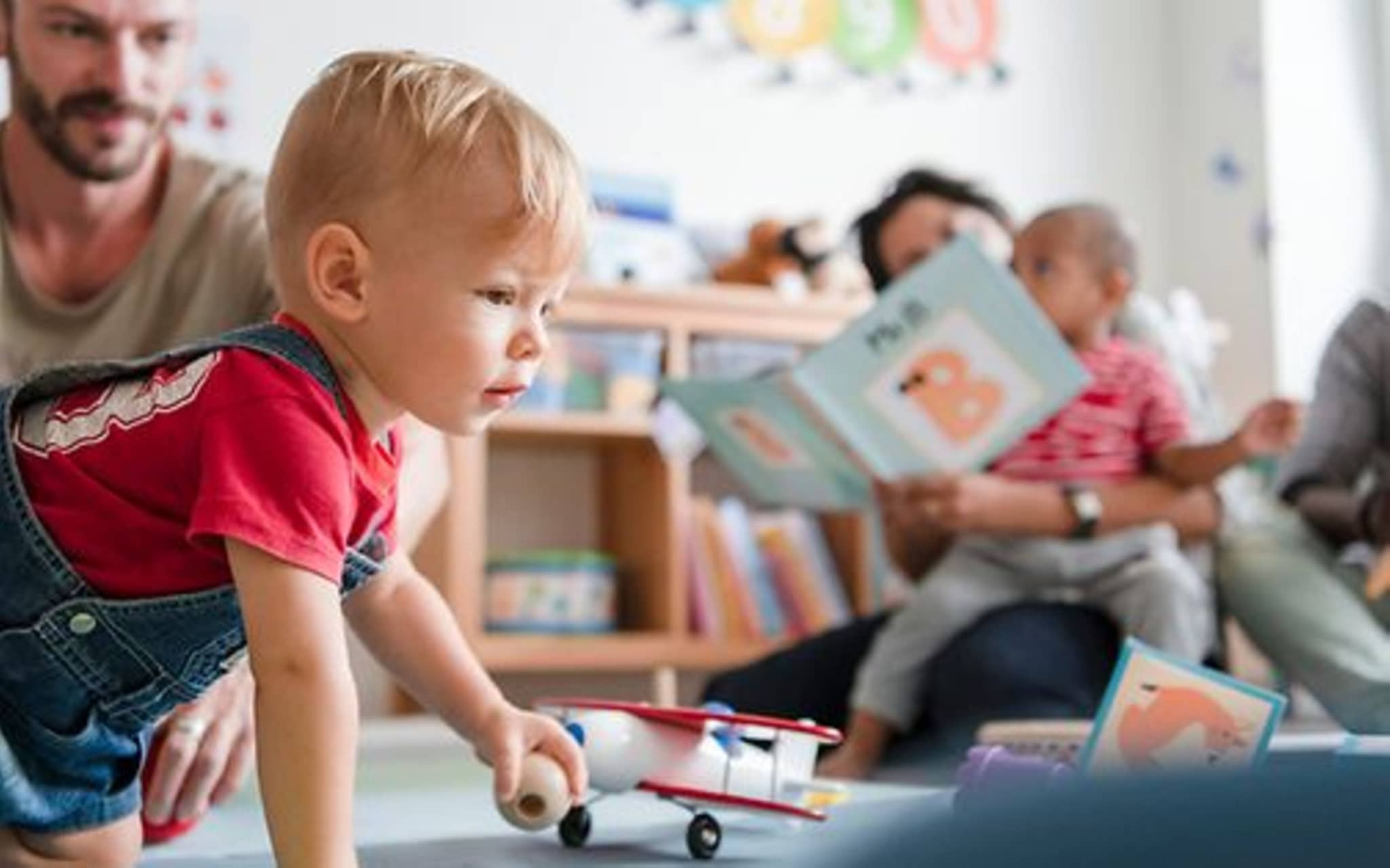 Symbolbild Kita-Eingewöhnung: Ein Vater sitzt mit seinem Kleinkind auf dem Fußboden einer Kita und beobachtet das Spielen