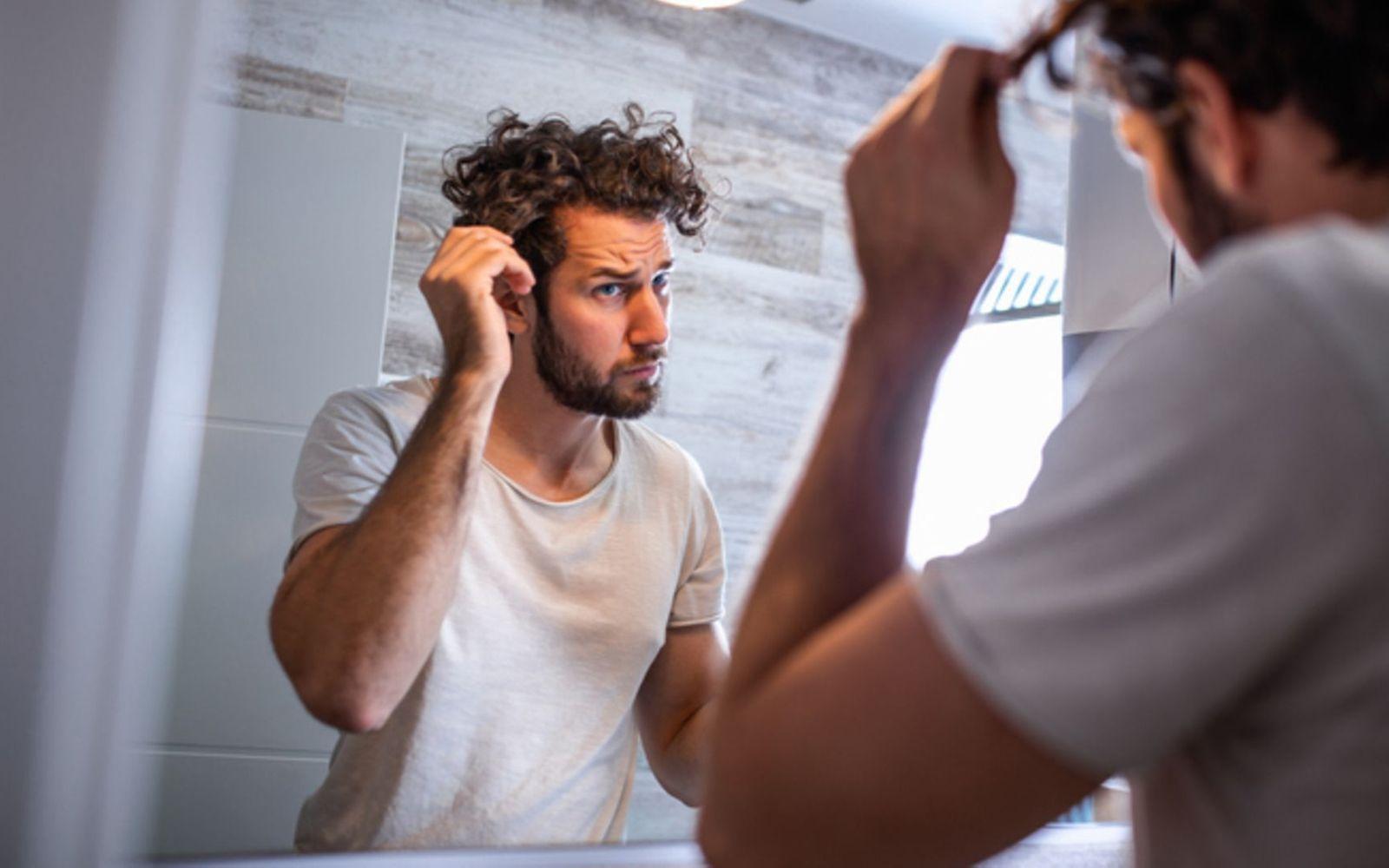 Haarausfall: Mann guckt skeptisch in den Spiegel und fasst sich ins Haar