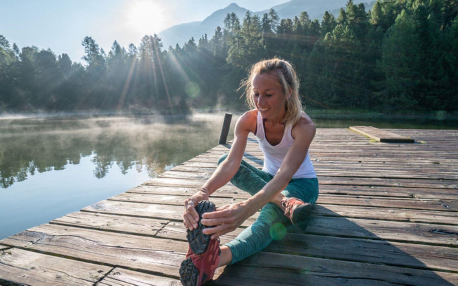 Laufen macht glücklich: Eine Frau sitzt auf einem Steg am See und stretcht sich.
