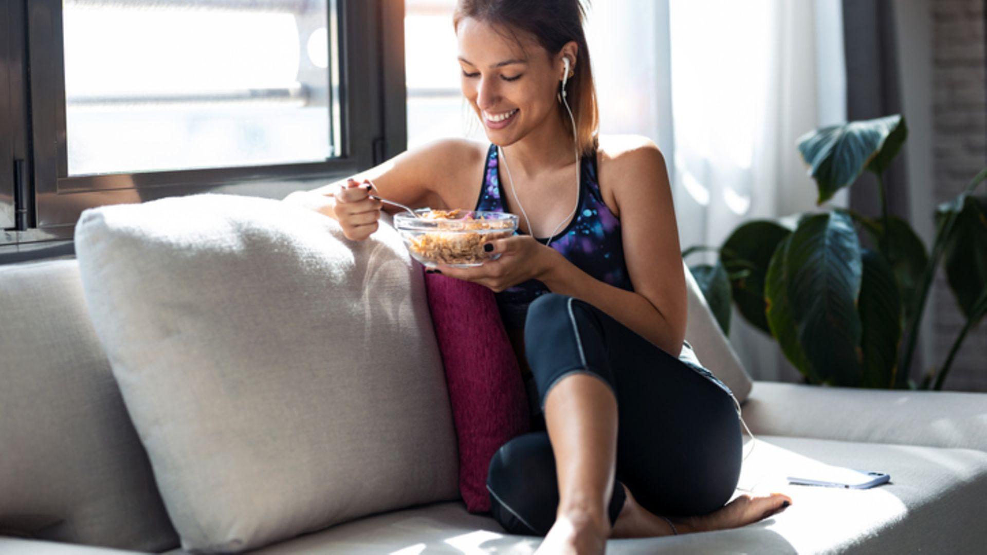 Ernährungs-Mythen: Junge Frau sitzt mit Sportklamotten auf einer Couch und isst eine Schüssel Müsli.