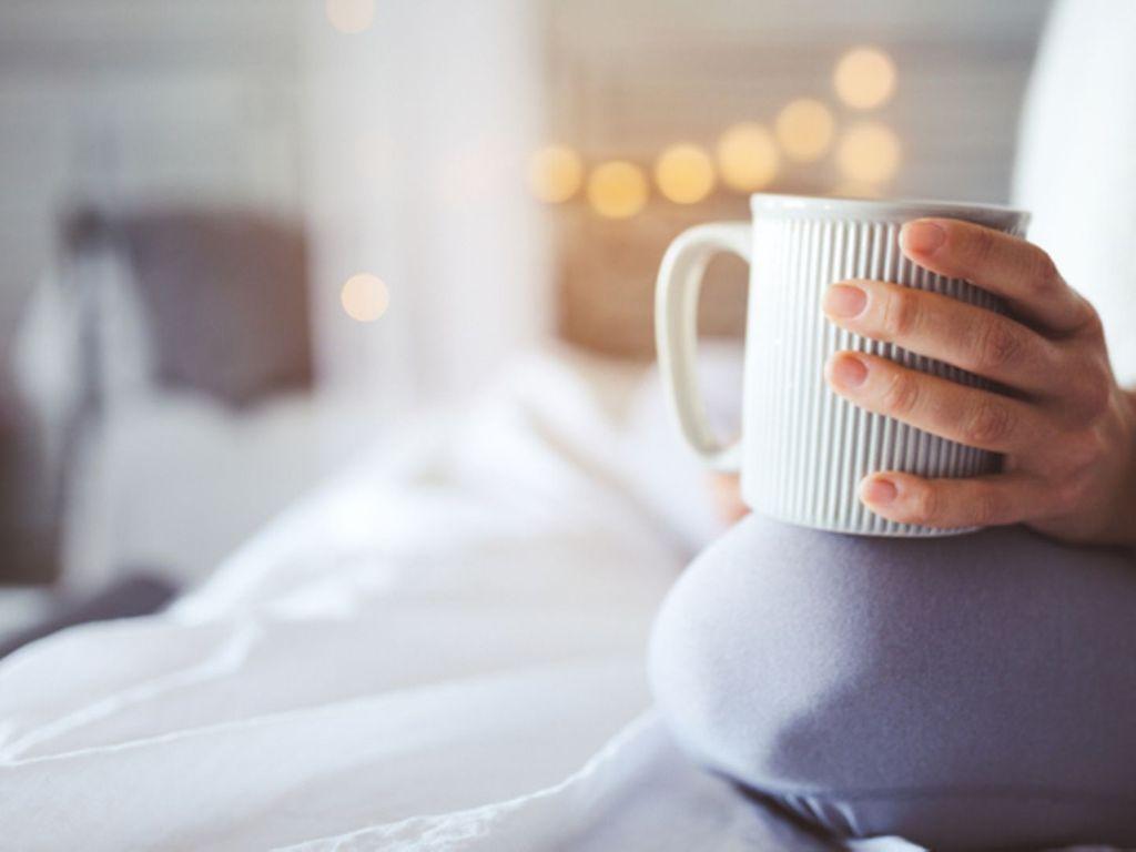 Morgenrituale: Eine Frau sitzt im Bett und hält eine Kaffeetasse in der Hand.