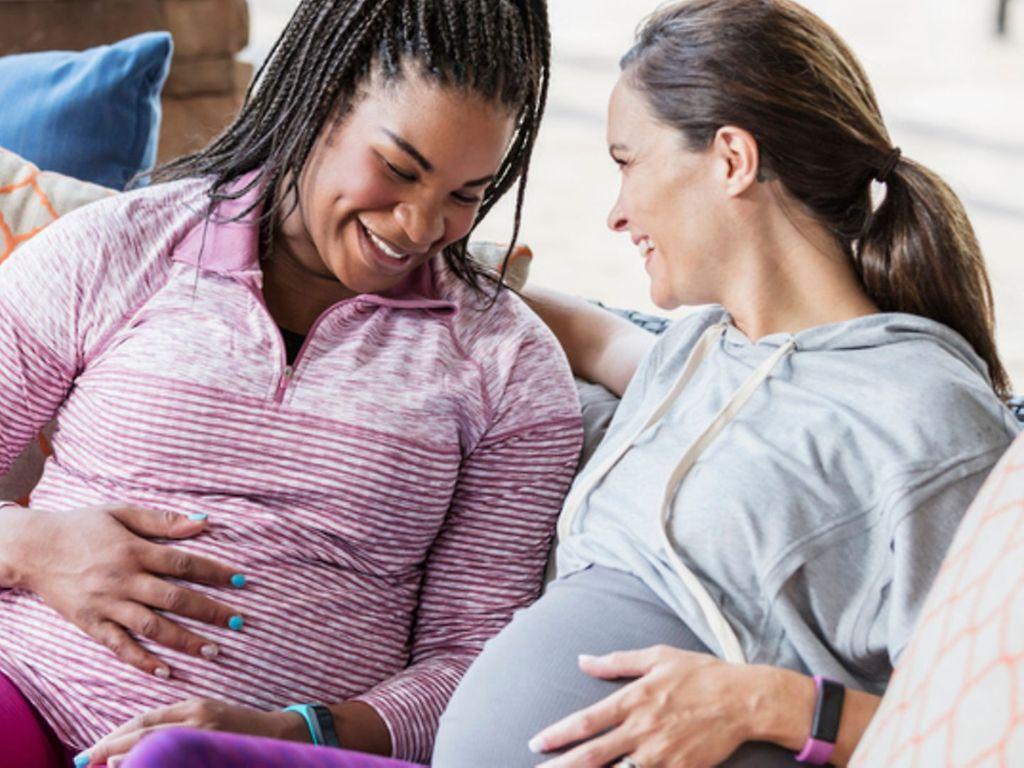 Fitnesstracker in der Schwangerschaft: Zwei Schwangere in Sportkleidung sitzen nebeneinander auf der Couch
