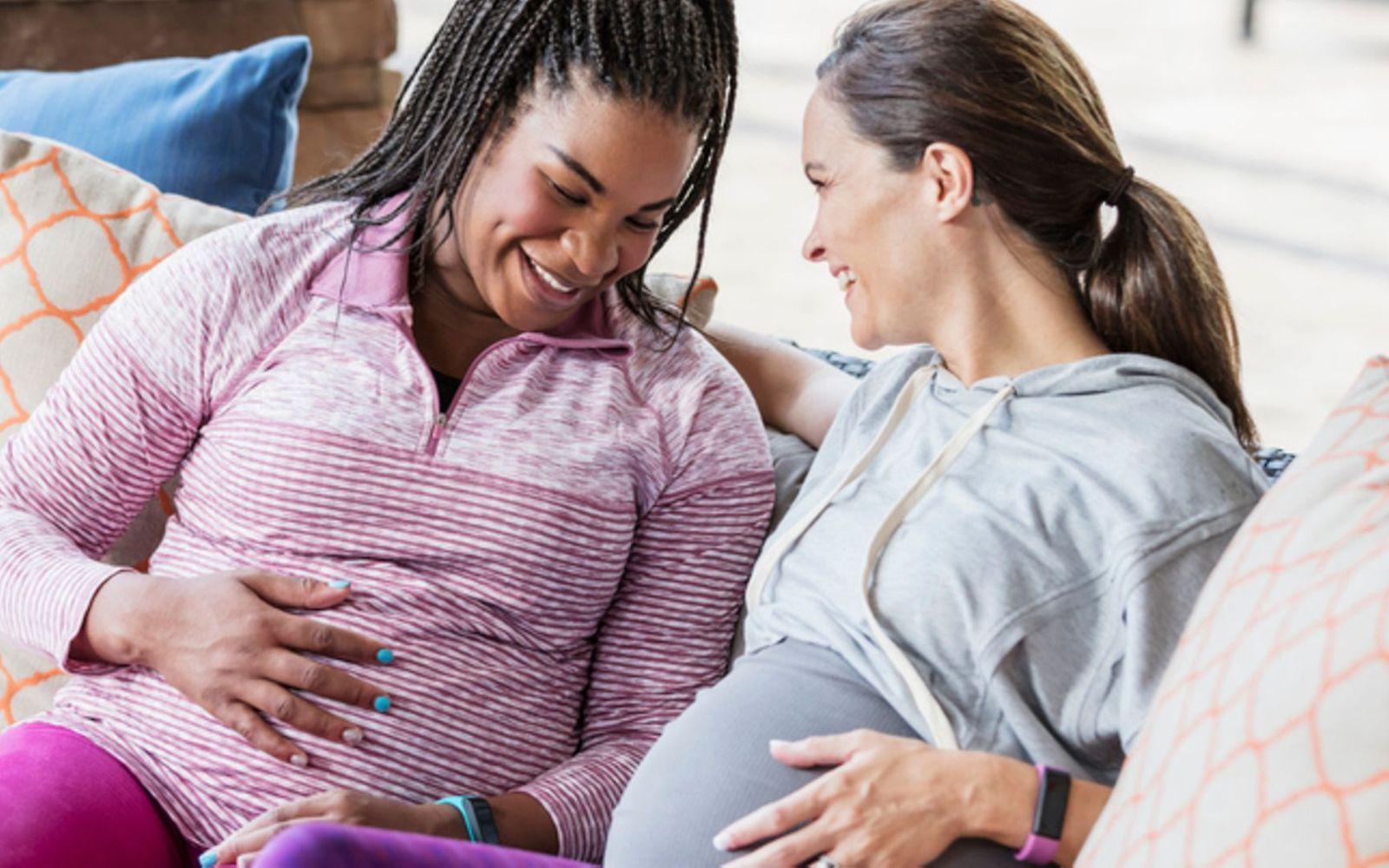 Fitnesstracker in der Schwangerschaft: Zwei Schwangere in Sportkleidung sitzen nebeneinander auf der Couch