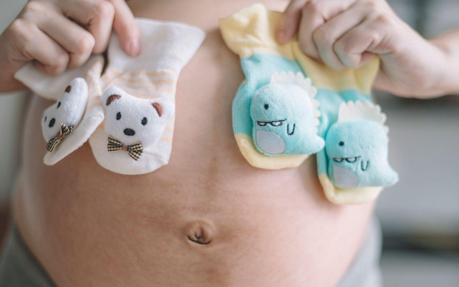 Zwillingsschwangerschaft: Frau hält Zwei Paar Söckchen vor ihren Schwangerschaftsbauch