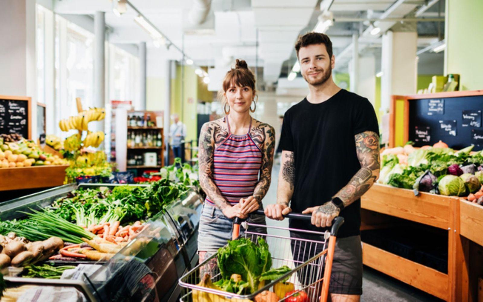 Vegane Ernährung und Nährstoffe: Junges Paar steht mit einem gefüllten Einkaufswagen in der Obst- und Gemüseabteilung.