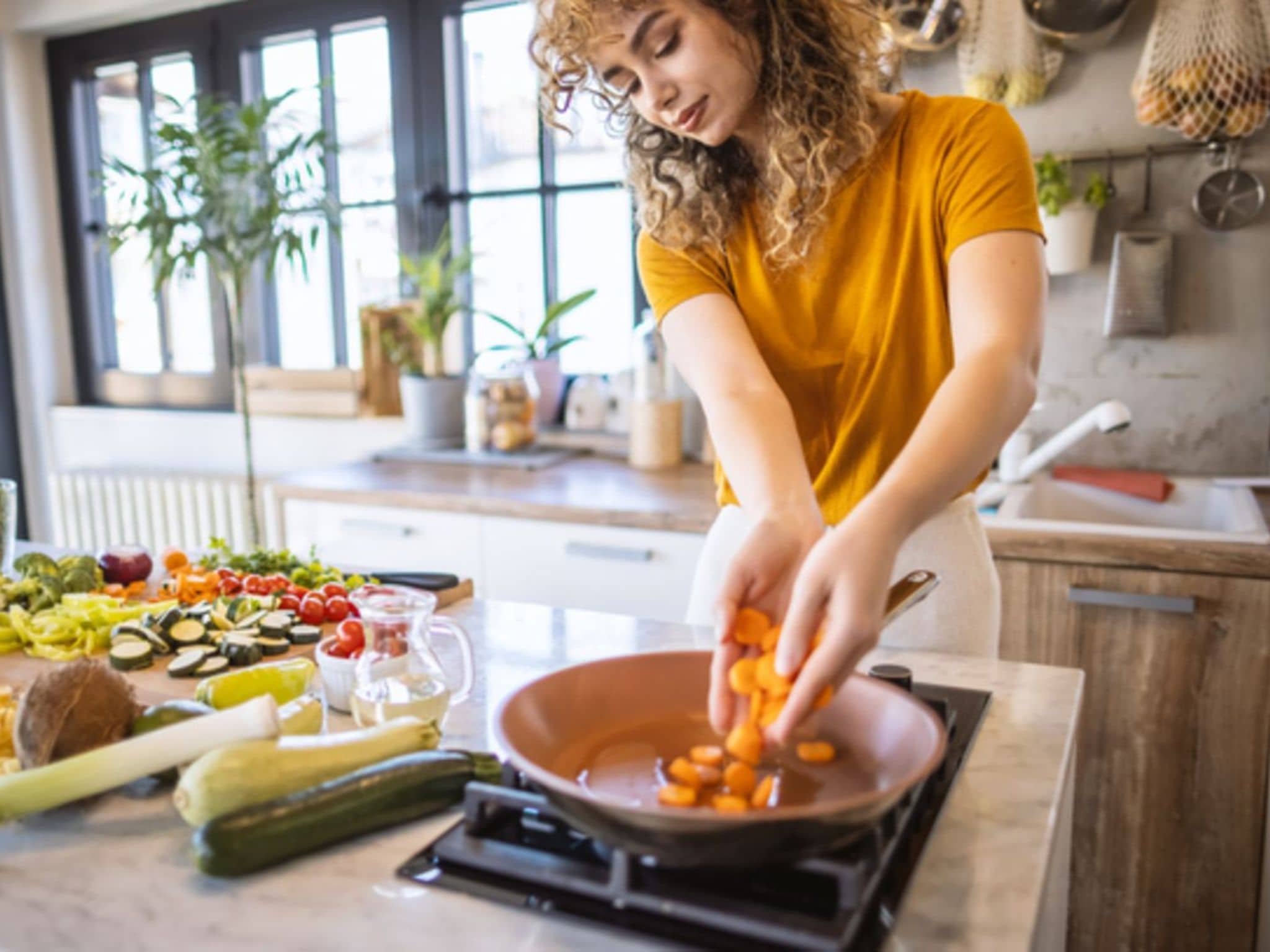 Intervallfasten: Junge Frau steht am Herd und schmeißt Karotten in eine Pfanne