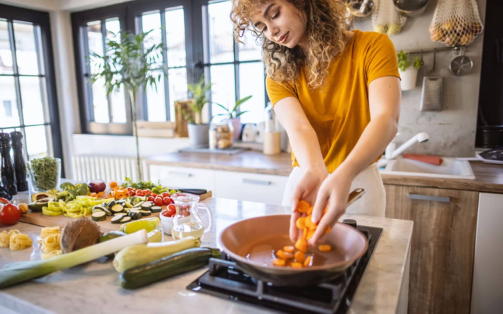Intervallfasten: Junge Frau steht am Herd und schmeißt Karotten in eine Pfanne
