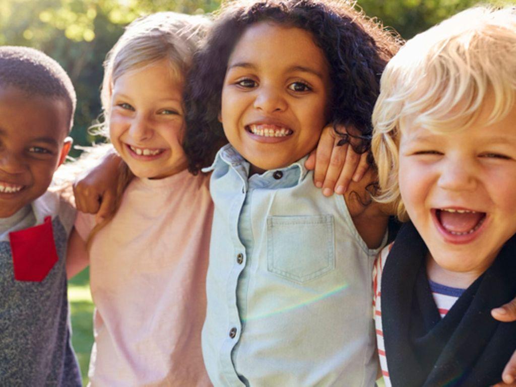 Tics bei Kindern: Vier kleine Kinder legen die Arme umeinander und lachen in die Kamera