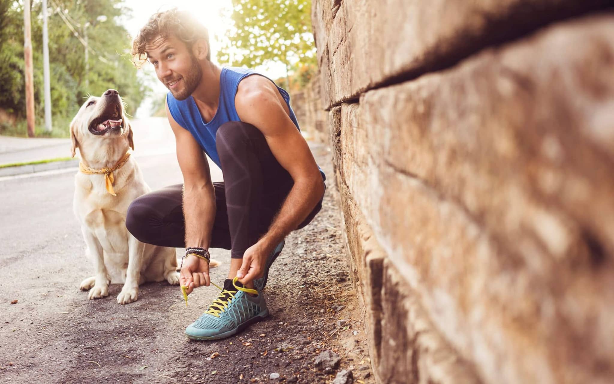 Schwitzender Mann macht sich kniend einen Laufschuh zu, daneben sitzt sein Hund.