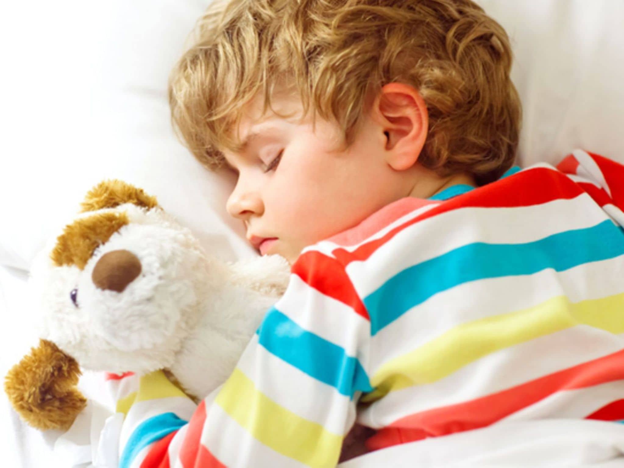 Bettnässen: Kleiner Junge liegt schlafend im Bett und hält seinen Kuschelhund im Arm.