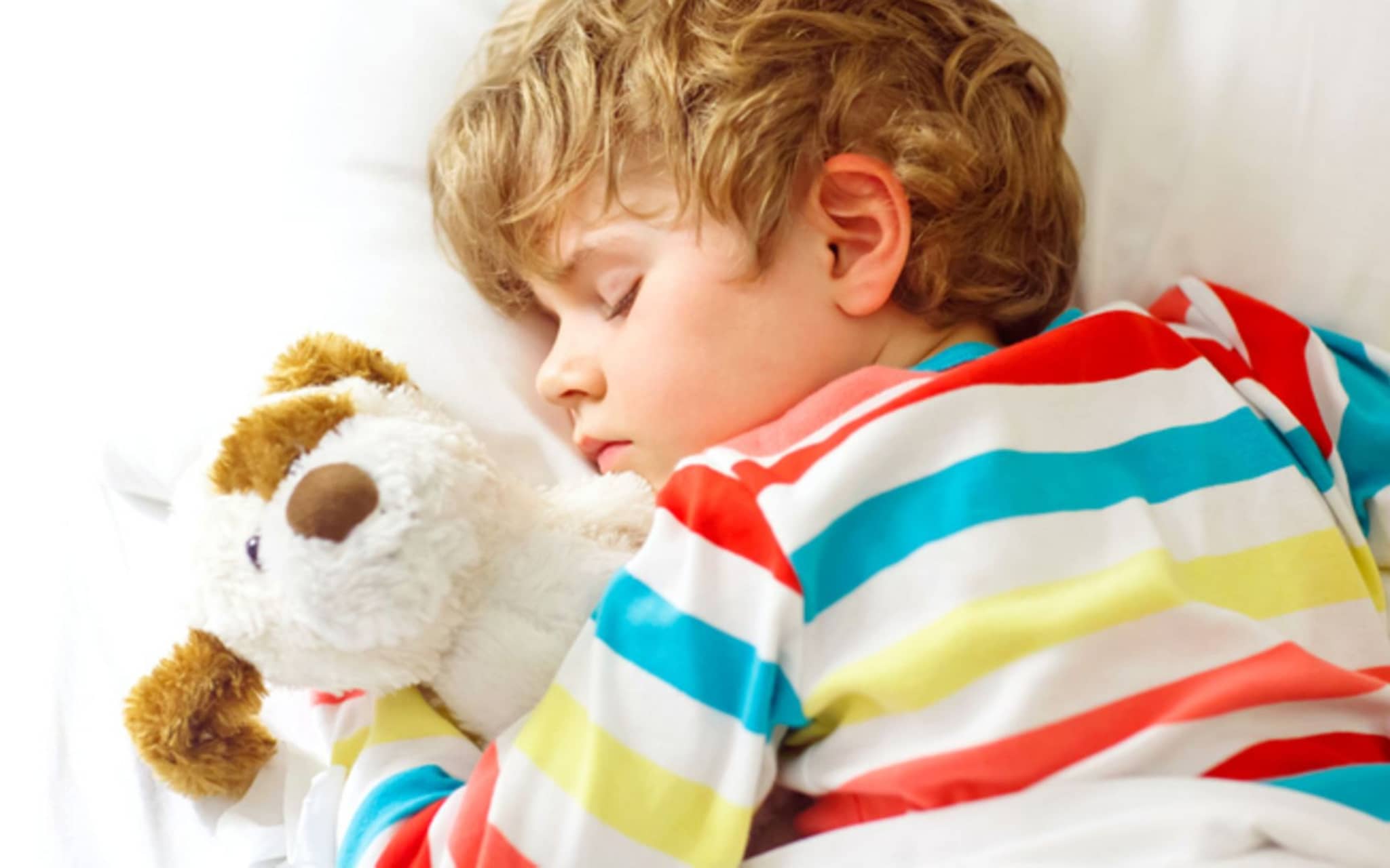Bettnässen: Kleiner Junge liegt schlafend im Bett und hält seinen Kuschelhund im Arm.