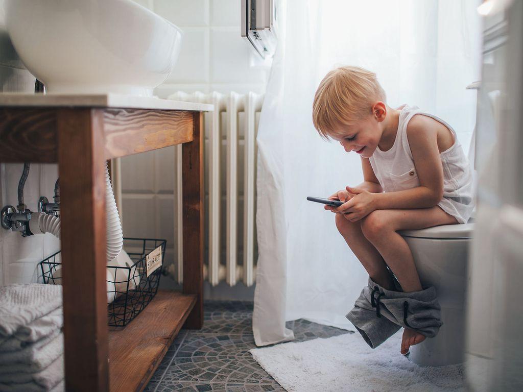 Verdauung und Darmerkrankungen: Junge mit Handy sitzt auf Toilette.