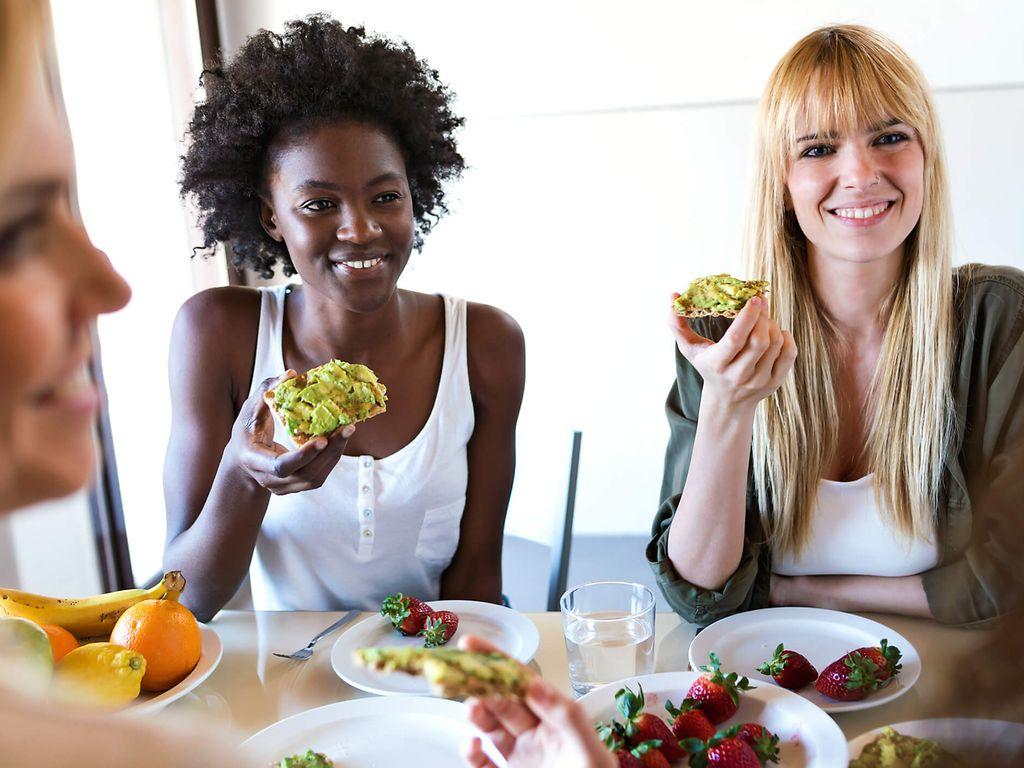 Drei junge Frauen, lachen und essen Brot mit Avocado zum Frühstück